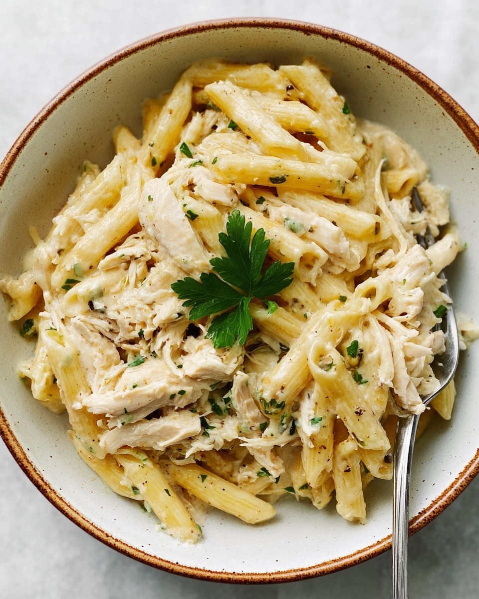 A bowl filled with creamy pasta made of pale yellow penne mixed with shredded light beige chicken in a white creamy sauce. Small bits of green herbs are scattered throughout the dish, adding a touch of color. On top, a small green parsley sprig sits in the center. A silver fork is partially inside the bowl on the right side, slightly lifting some pasta. The bowl is white with a rustic brown rim, sitting on a white marbled surface. photo taken with an iphone --ar 4:5 --v 7