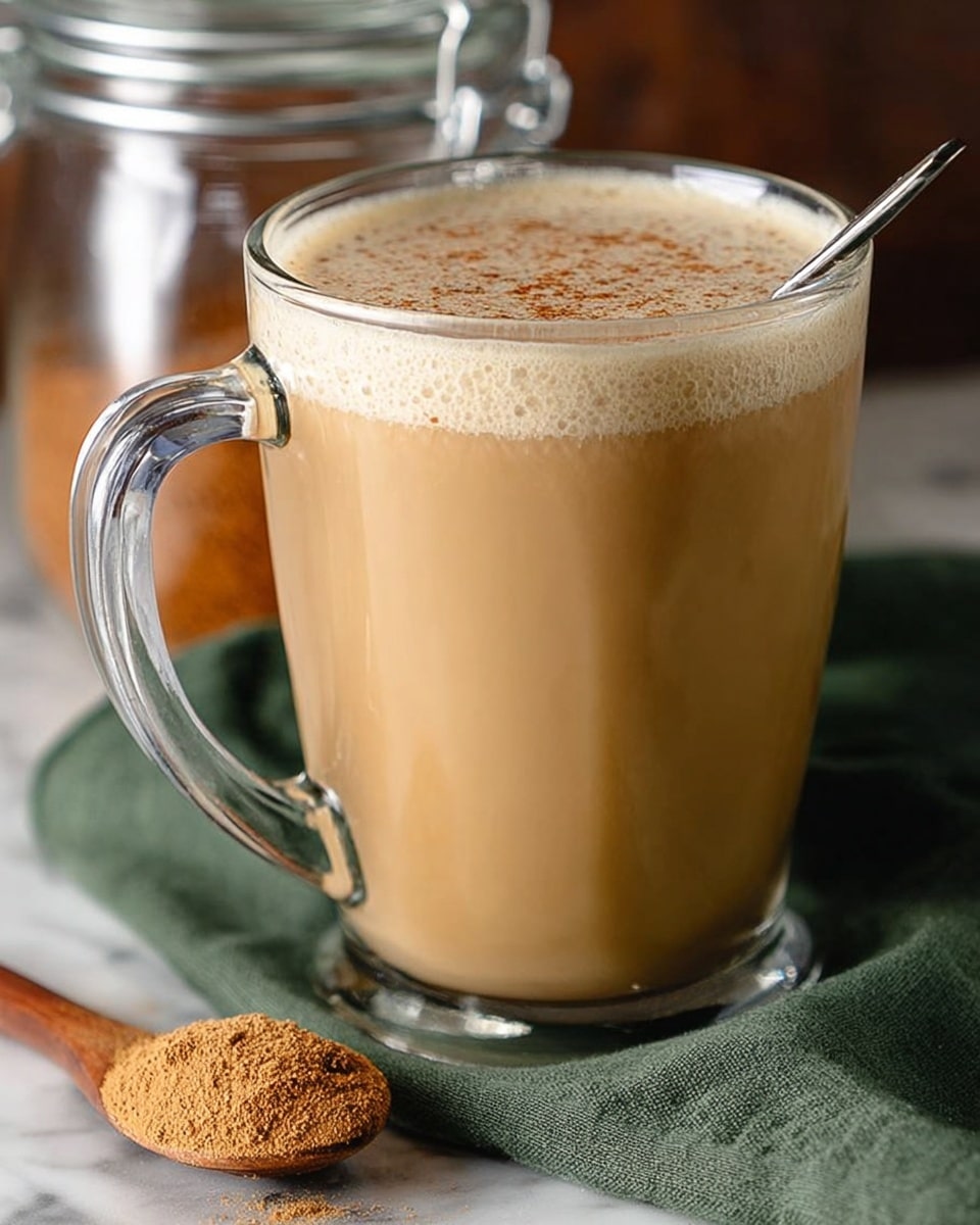 A clear glass mug filled with creamy tan chai tea, topped with a light layer of foam sprinkled with cinnamon powder. The mug has a thick handle on the left side and a silver spoon placed inside, leaning against the right edge. The mug sits on a white marbled surface with a green cloth underneath, and a wooden spoon with powdered chai is beside it. In the background, there is a blurry glass jar with a latch. photo taken with an iphone --ar 4:5 --v 7