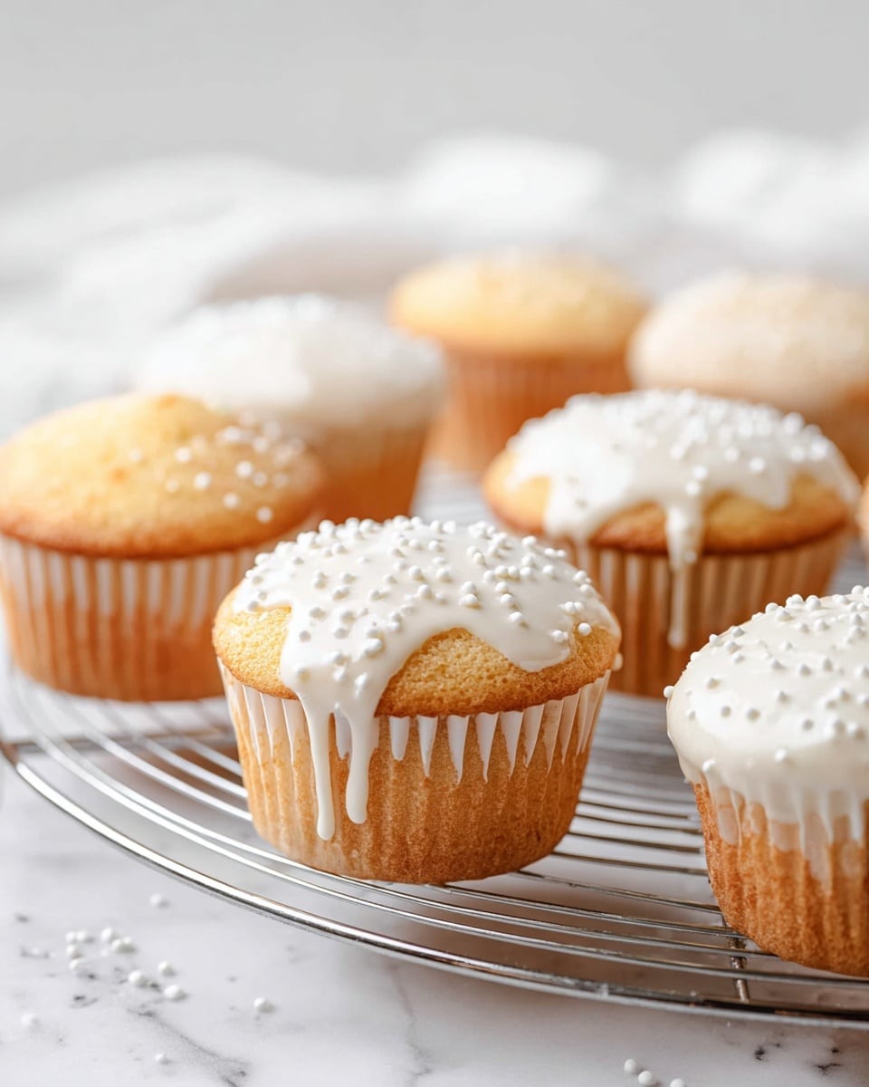 The image shows a group of cupcakes sitting on a round wire cooling rack over a white marbled surface. Each cupcake has one layer of light golden brown cake with a slightly rough texture on top. Some cupcakes are topped with smooth white icing dripping slightly down the sides, decorated with small white round sprinkles. Other cupcakes have no icing and have a shiny sugar glaze sprinkled over their tops. The cupcake liners are light brown and white. The scene is bright and clean looking with soft natural light. photo taken with an iphone --ar 4:5 --v 7