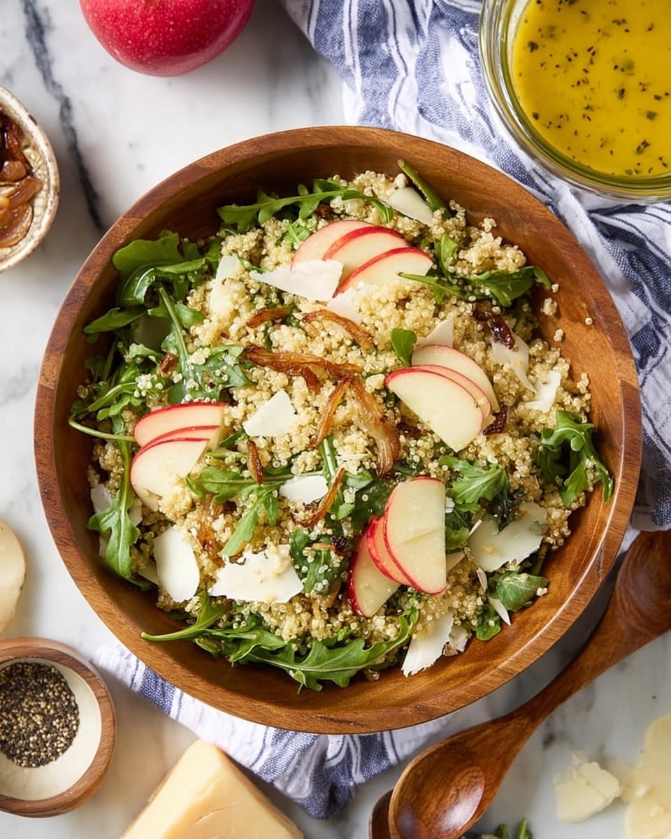 In a deep wooden bowl, a mixed salad shows three main layers: at the base, light beige quinoa forms a grainy texture; scattered on top are thin slices of red and white apple adding smooth, curved shapes; bright green arugula leaves spread throughout bring a fresh look with their jagged edges. Small white shavings of cheese are sprinkled over the salad, adding soft patches, while toasted shallots give a crispy, golden-brown touch all around. The bowl rests partly on a white and blue striped cloth over a white marbled surface, with a wooden spoon beside it and a glass bowl of yellow dressing with black pepper in the background. A red apple and a piece of pale cheese are also nearby. photo taken with an iphone --ar 4:5 --v 7