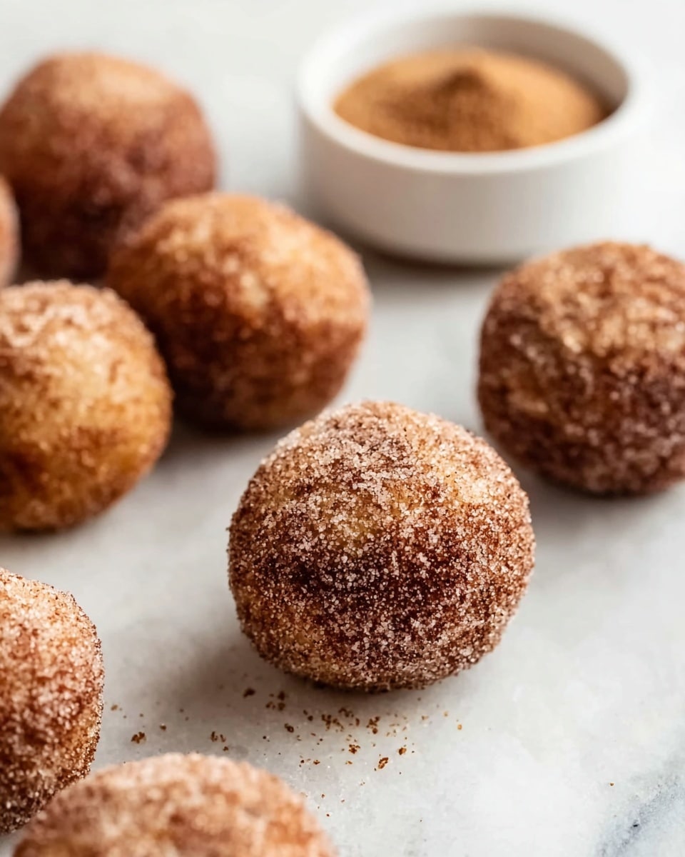 The image shows several round dough balls covered in a dark brown cinnamon sugar coating, placed on a white marbled surface. The dough balls have a rough texture from the sugar and cinnamon layer around them. A small white bowl filled with more cinnamon sugar sits to the side, slightly out of focus. The round dough balls are arranged randomly on the surface, with one in the foreground in full focus and others blurred in the background. The overall color palette of the dish is warm brown shades with the white bowl and marbled surface creating a clean background. photo taken with an iphone --ar 4:5 --v 7