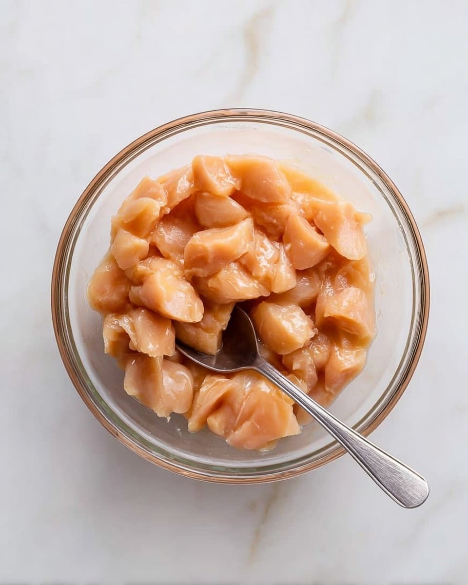 A clear glass bowl sits on a white marbled surface, filled with evenly cut small pieces of raw chicken that have a pale pink and light orange color. A silver spoon rests inside the bowl, partially covered by the chicken pieces. The bowl's contents look fresh and smooth, showing slight shine from moisture. Photo taken with an iphone --ar 4:5 --v 7