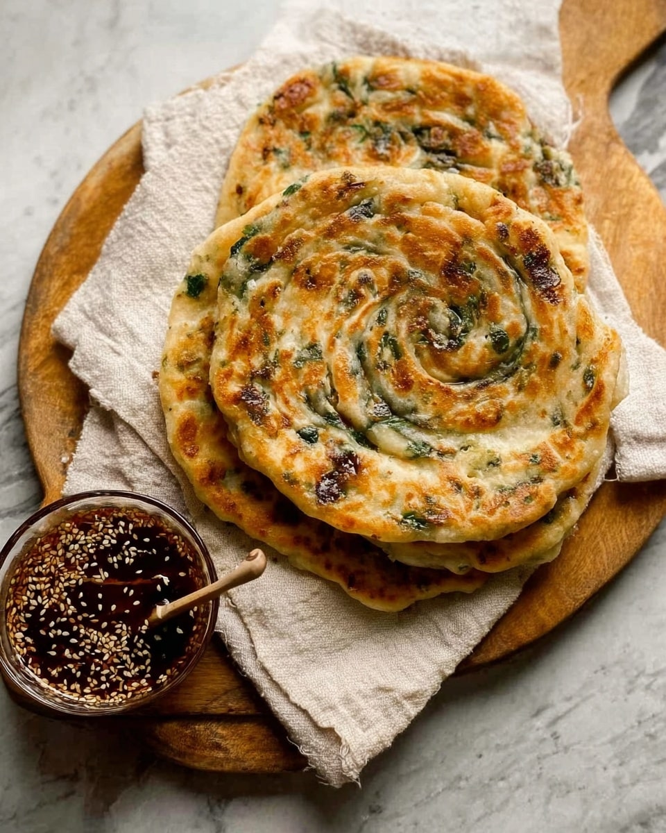 The image shows three round flatbreads stacked on a wooden board that rests on a cream-colored cloth. Each flatbread has a golden-brown and light beige spiral pattern with green spots scattered throughout, indicating some kind of herb or vegetable inside the dough. Next to the flatbreads, there is a small glass bowl filled with dark brown sauce topped with white sesame seeds, and a wooden spoon is resting inside the bowl. All of this is set on a white marbled surface. photo taken with an iphone --ar 4:5 --v 7