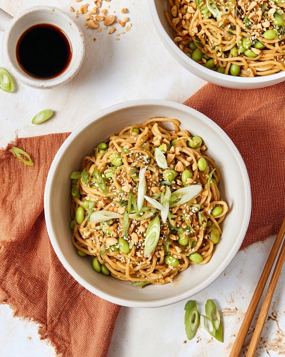 The dish shows two white bowls filled with a noodle salad placed on a white marbled surface. Each bowl holds a mix of light brown noodles tangled with bright green edamame beans, small chopped peanuts, and scattered white sesame seeds. Thin slices of fresh green onions are sprinkled on top, adding a fresh contrast to the warm noodles. Near the bowls, there is a small white cup with dark soy sauce and a pair of wooden chopsticks. The background has a textured orange cloth under the main bowl, with some sesame seeds and green onions casually placed nearby. photo taken with an iphone --ar 4:5 --v 7