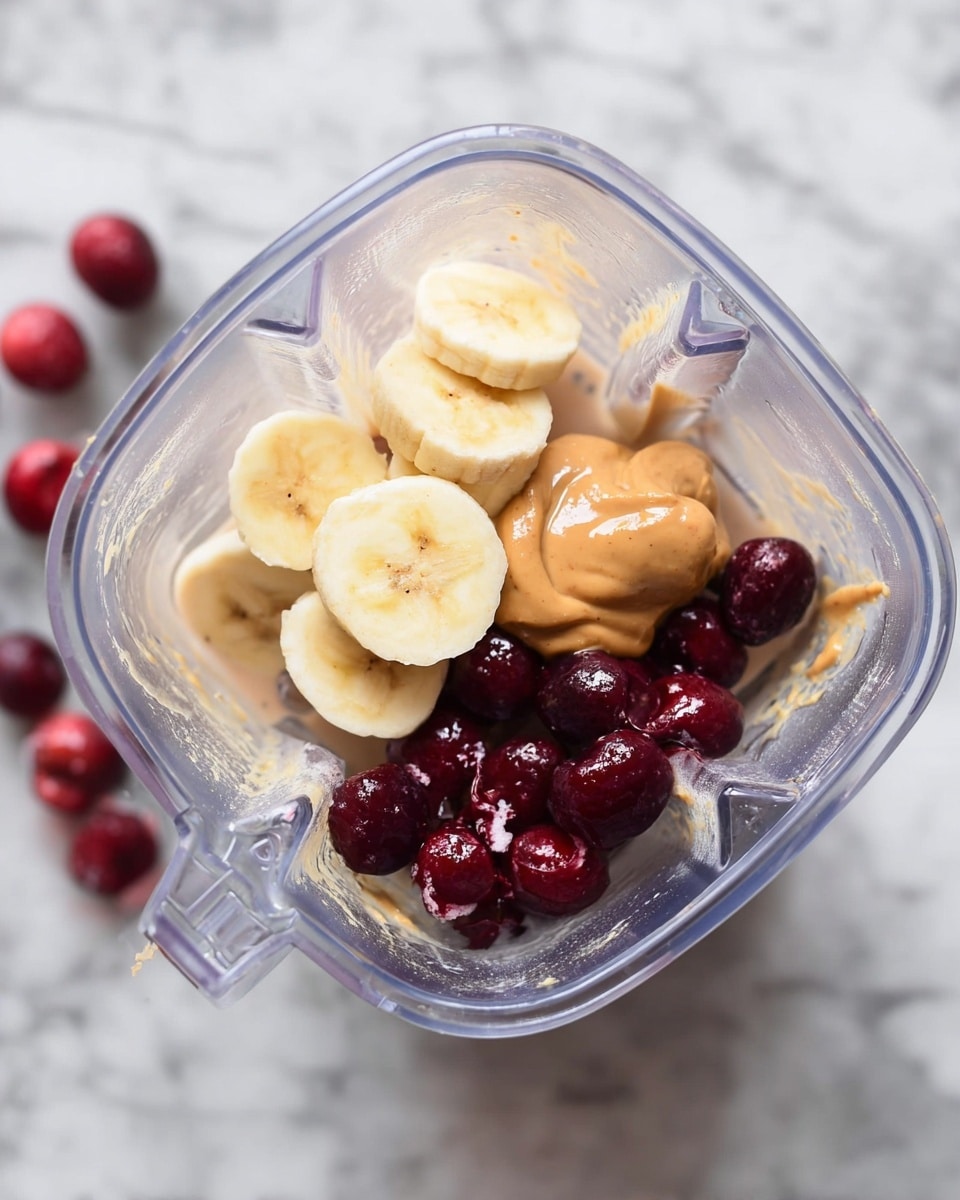Inside a clear blender, there are four layers of ingredients visible from top view: on the left, three thick slices of yellow banana with soft texture; next to it on the right is a smooth, light brown dollop of creamy peanut butter; below the banana and peanut butter are deep red and shiny whole cherries, some split open showing their juicy inside; and underneath all of this is a light beige creamy liquid, likely milk, pooling at the bottom. The blender is placed on a white marbled surface, with a few loose cherries scattered nearby. photo taken with an iphone --ar 4:5 --v 7