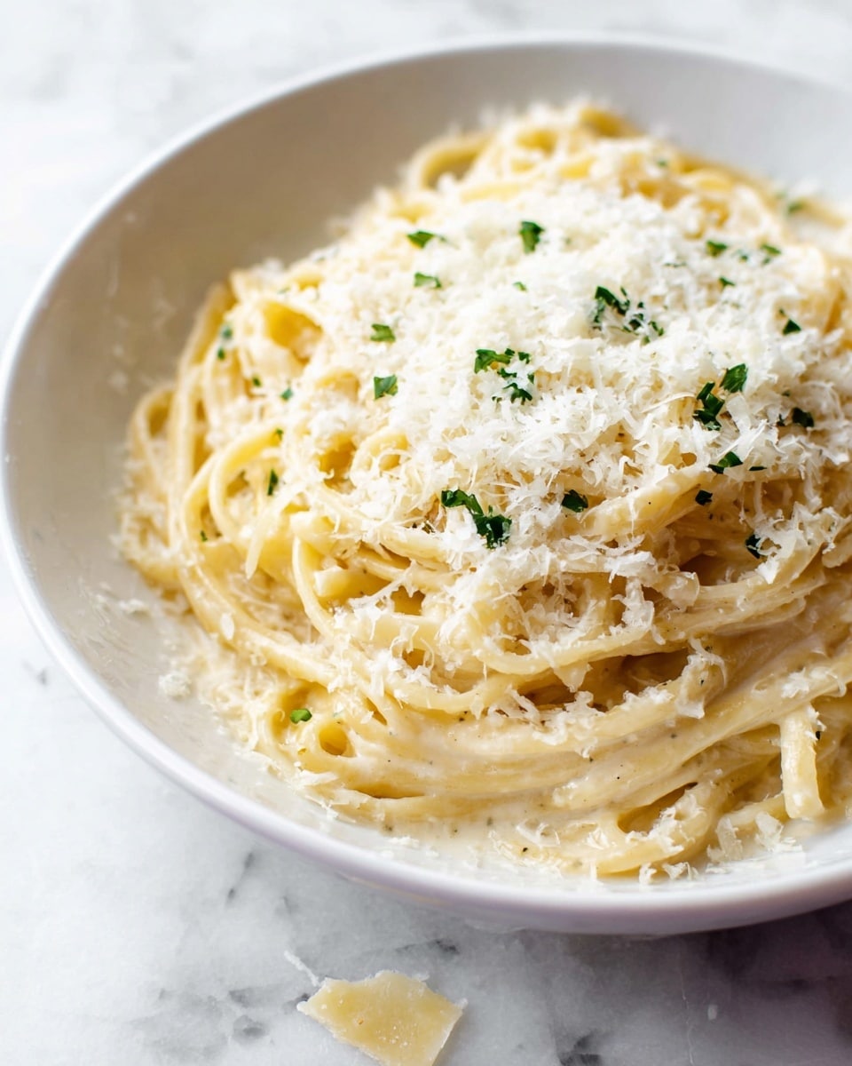 A close-up view of a white bowl filled with a single layer of creamy linguine pasta. The pasta is coated evenly with a smooth, light beige sauce and topped with a generous layer of finely grated white cheese that looks fluffy and soft. Small bits of bright green herbs are scattered sparingly over the cheese, adding color contrast. The bowl is placed on a white marbled surface, and a few cheese shavings have fallen around it. The overall look is warm, creamy, and inviting. Photo taken with an iphone --ar 4:5 --v 7