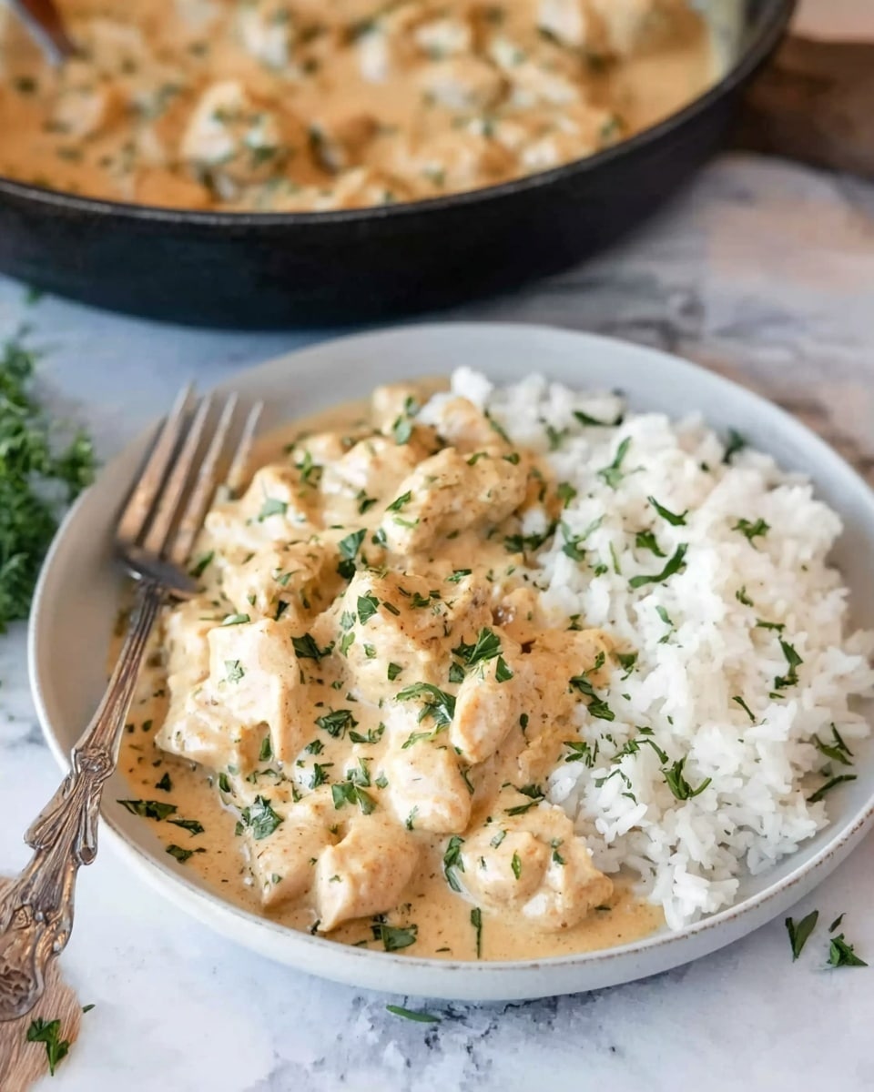 A white round plate filled with two main layers: at the bottom, a textured layer of fluffy white rice; on top, a creamy light beige sauce with small pieces of chicken mixed throughout, sprinkled with small green herbs as garnish. A fork is placed on the left side of the plate. In the background, a black pan holds more creamy chicken dish with the same light beige sauce and green herbs visible. The surface is a white marbled texture. photo taken with an iphone --ar 4:5 --v 7