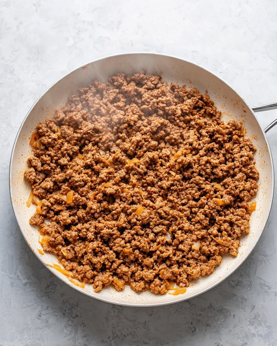 A large white skillet filled with cooked ground meat that is brown and crumbly, mixed with small soft pieces of cooked onion that are light yellow and slightly translucent. The meat looks seasoned with a reddish spice coating its uneven surface. Steam rises gently from the skillet. The skillet is placed on a white marbled surface. photo taken with an iphone --ar 4:5 --v 7