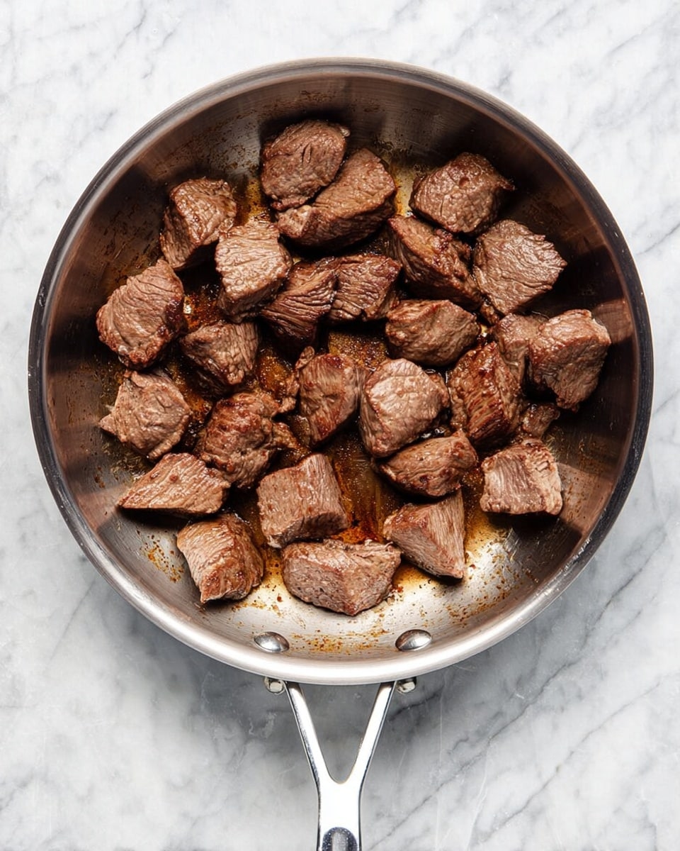 A stainless steel pan filled with several medium-sized chunks of cooked brown meat sits on a white marbled surface. The meat pieces vary in size and shape, some showing cooked texture with slight browning and visible grain, while the pan has some browned spots where the meat has cooked. The pan features a handle on one side and a loop handle on the other, both shiny and metallic. photo taken with an iphone --ar 4:5 --v 7