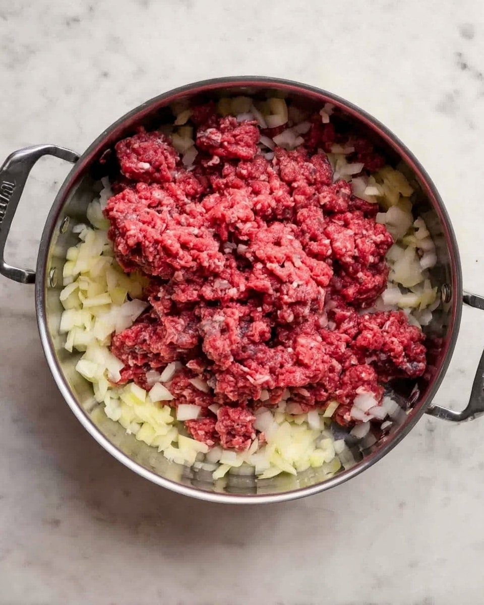 A round metal pan with two handles contains a base layer of diced onions, pale yellow and slightly translucent, spread evenly covering the bottom. Above the onions, raw ground beef with a dense, chunky texture is scattered unevenly, showing a bright red color mixed with some pale pink and grey spots. The pan sits on a flat surface of white marbled texture. The whole scene is simple and uncooked. Photo taken with an iphone --ar 4:5 --v 7