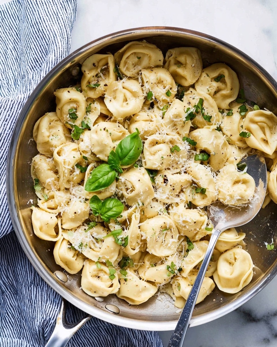 A shiny silver pan filled with creamy tortellini pasta scattered evenly, topped with small green basil leaves and finely grated white cheese. The pasta pieces are light yellow with a soft texture mixed with light seasoning spots and a silver spoon resting on the right side inside the pan. The pan is placed on a white marbled surface with a folded blue and white striped cloth on the top left corner. photo taken with an iphone --ar 4:5 --v 7