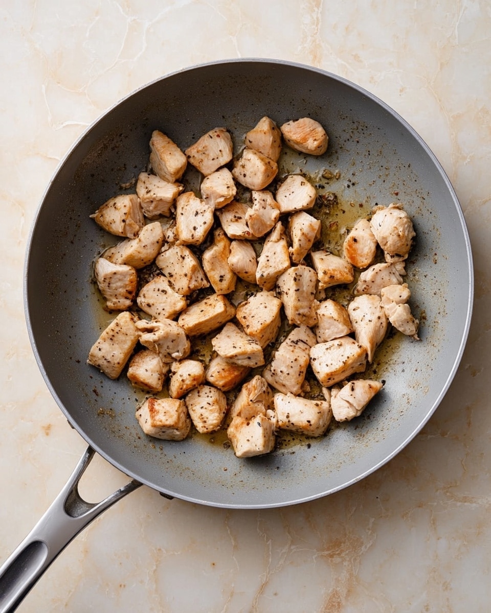 A gray pan filled with small cooked chicken pieces that are light brown with some black pepper spots, spread evenly inside the pan showing a slightly shiny and oily texture on the surface. The pan handle is long and metallic, resting on a white marbled surface with light beige patterns. Photo taken with an iphone --ar 4:5 --v 7