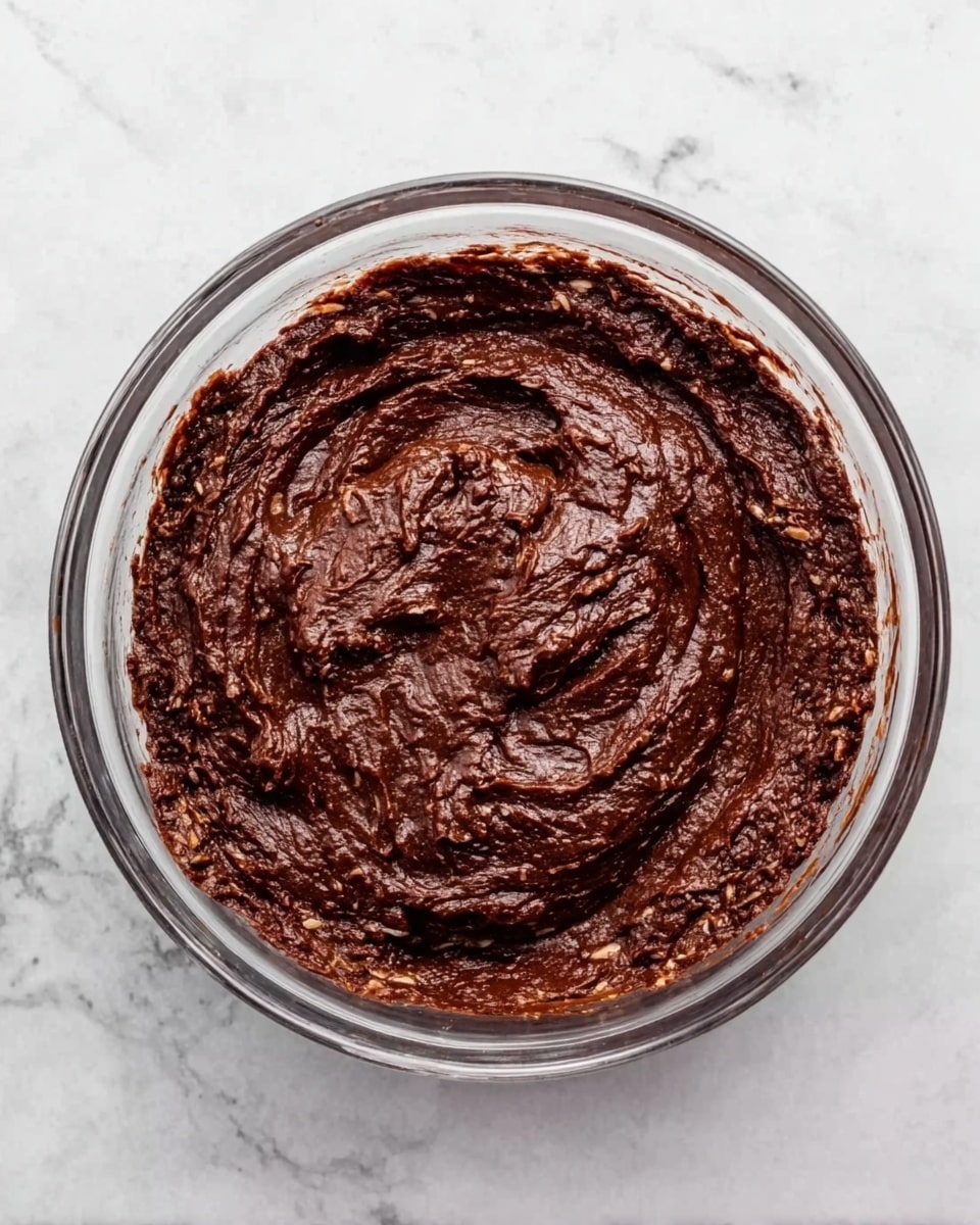 A clear glass bowl is filled with a dark brown, thick mixture that looks slightly chunky with visible bits of lighter brown or beige. The bowl sits on a white marbled surface, and the texture of the mixture looks dense and moist, swirling slightly on top as if just stirred. The overall appearance suggests a rich, chocolatey batter or paste. photo taken with an iphone --ar 4:5 --v 7