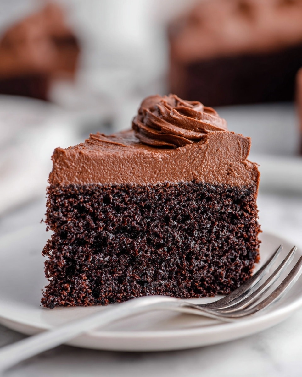 A close-up view of a chocolate cake slice on a white plate, showing two clear layers: the bottom layer is a dark, dense, and moist chocolate cake with a slightly crumbly texture, while the top layer is a smooth, thick chocolate frosting with a creamy, whipped texture and soft peaks. The background features a soft blur of similar cake slices on a white marbled surface, with a white fork placed next to the plate. photo taken with an iphone --ar 4:5 --v 7