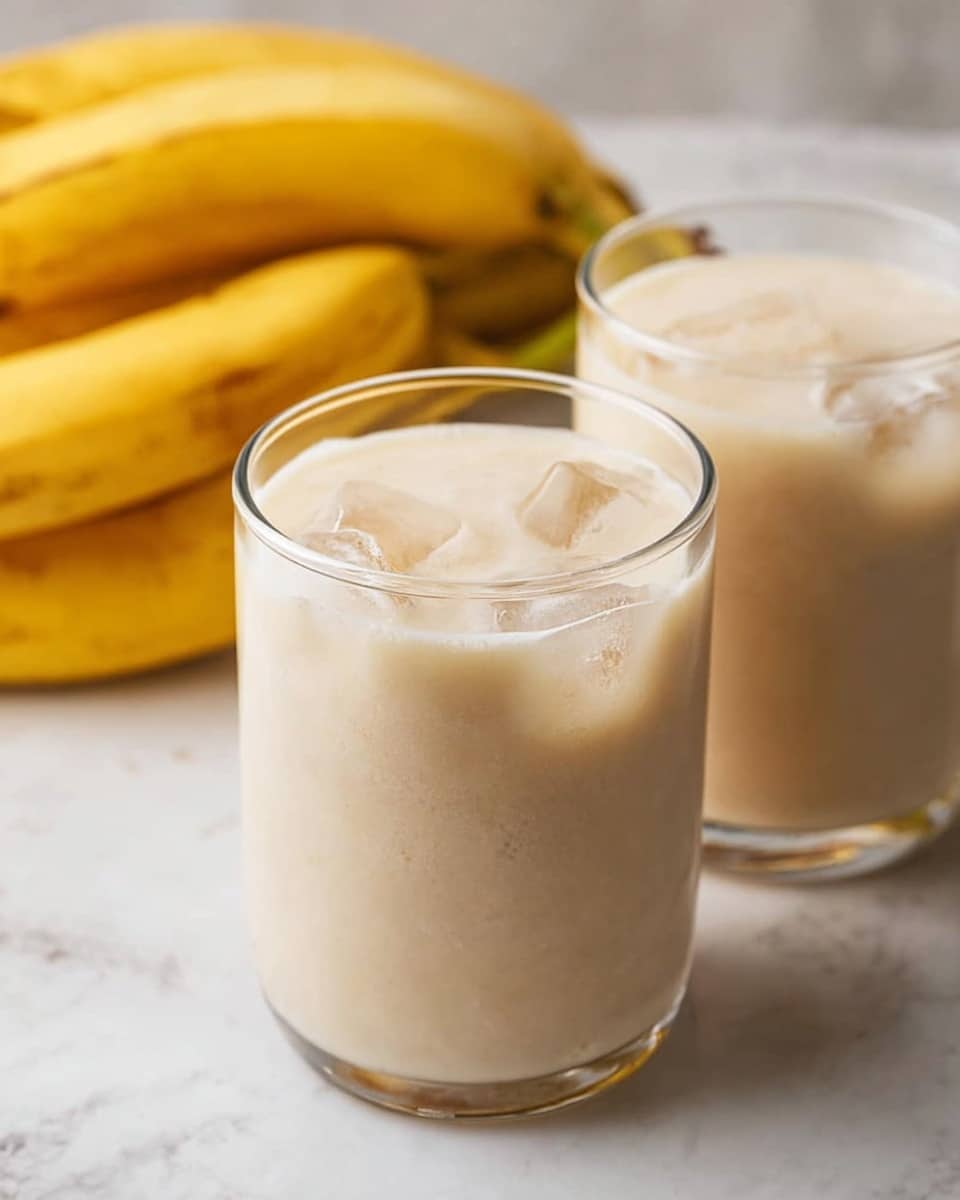 Two clear glasses filled with a light creamy beige banana smoothie, with visible ice cubes floating near the creamy top layer. Behind the glasses, there are ripe yellow bananas resting on a white marbled surface. The scene is bright and clean with soft natural light highlighting the smooth texture of the smoothie and the fresh bananas. The background is softly blurred to keep focus on the glasses and the bananas. photo taken with an iphone --ar 4:5 --v 7