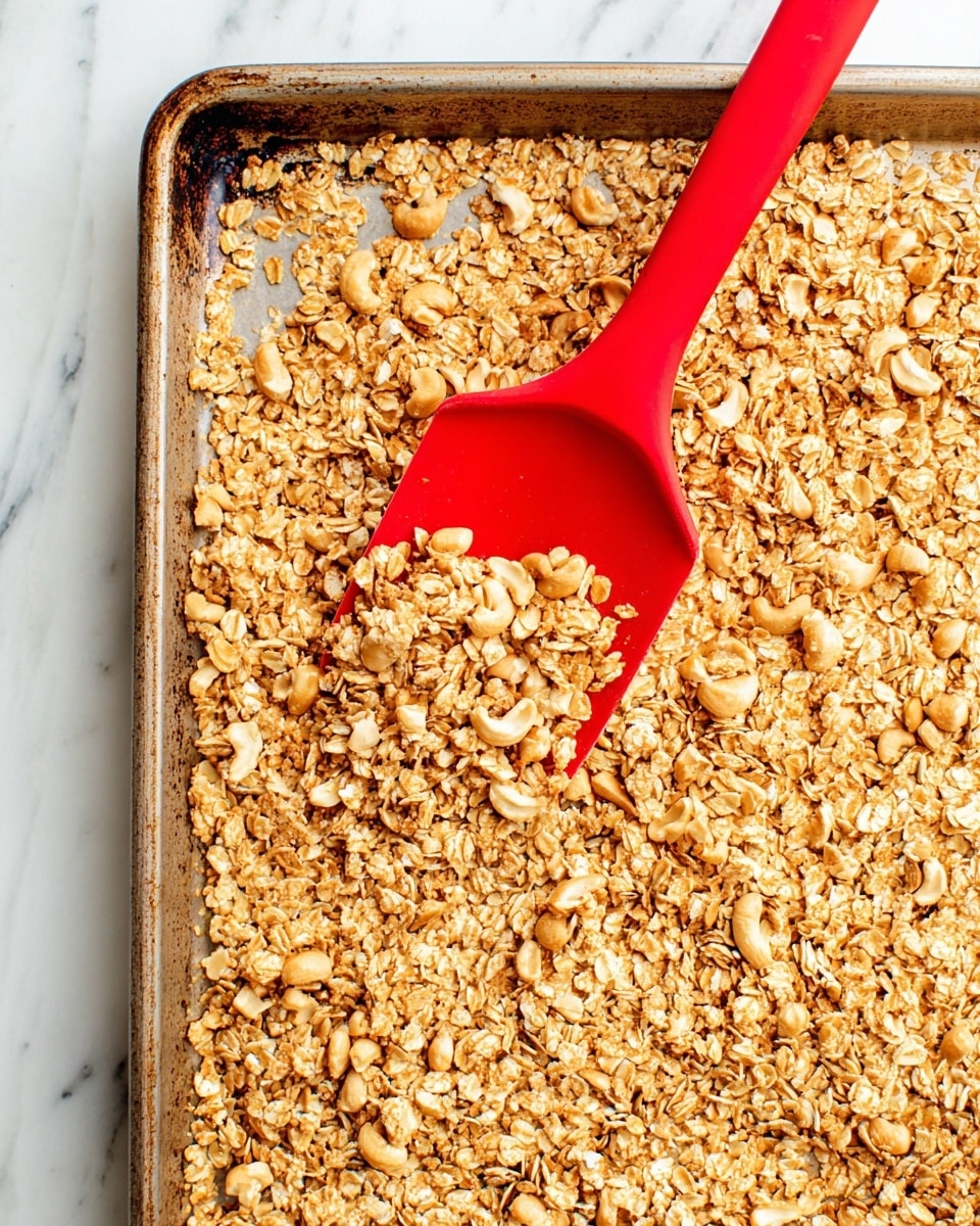 The image shows a close-up of a baking tray filled with a layer of golden brown oats mixed with cashew pieces, giving the texture a crunchy, rough look. In the middle of the tray, there is a bright red spatula lifting a portion of the oat mix, with the oats clinging to it. The tray has a slightly browned, used appearance on its edges. The background surface is a white marbled texture. photo taken with an iphone --ar 4:5 --v 7