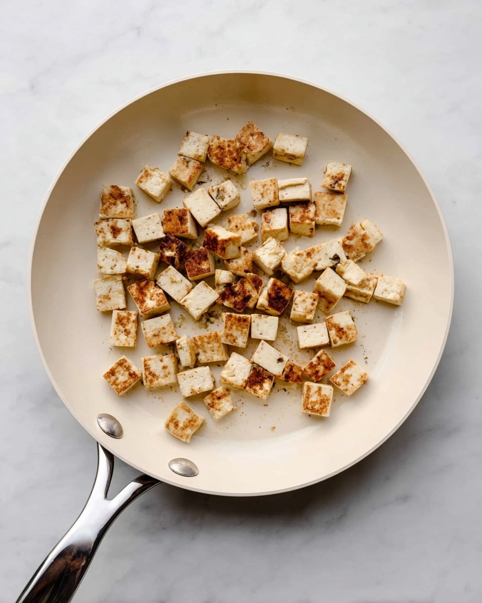 A white pan with a silver handle holds small, cubed pieces of tofu that are cooked to a light brown with some crispy edges. The tofu pieces are spread unevenly in the middle of the pan, showing a mix of golden and off-white colors with a soft texture. The background is a clean white marbled surface. photo taken with an iphone --ar 4:5 --v 7