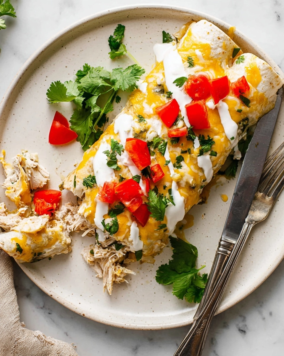 A white plate holds a rolled enchilada dish topped with melted yellow and white cheese, white creamy sauce drizzled on top, and small bright red tomato pieces scattered around. Fresh green cilantro leaves are placed both on and beside the enchiladas. On the left side, a piece of the enchilada has been pulled apart, showing shredded white meat inside. A silver fork holds a small red tomato piece on the edge of the plate, and a silver knife rests on the right side. The background is a white marbled surface. photo taken with an iphone --ar 4:5 --v 7