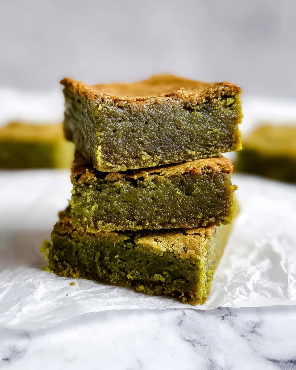 A stack of three square green brownies with a moist, dense texture and a golden-brown thin crust on the top layer, all placed on a wrinkled white paper over a white marbled surface, with two additional green brownie pieces blurred in the background photo taken with an iphone --ar 4:5 --v 7