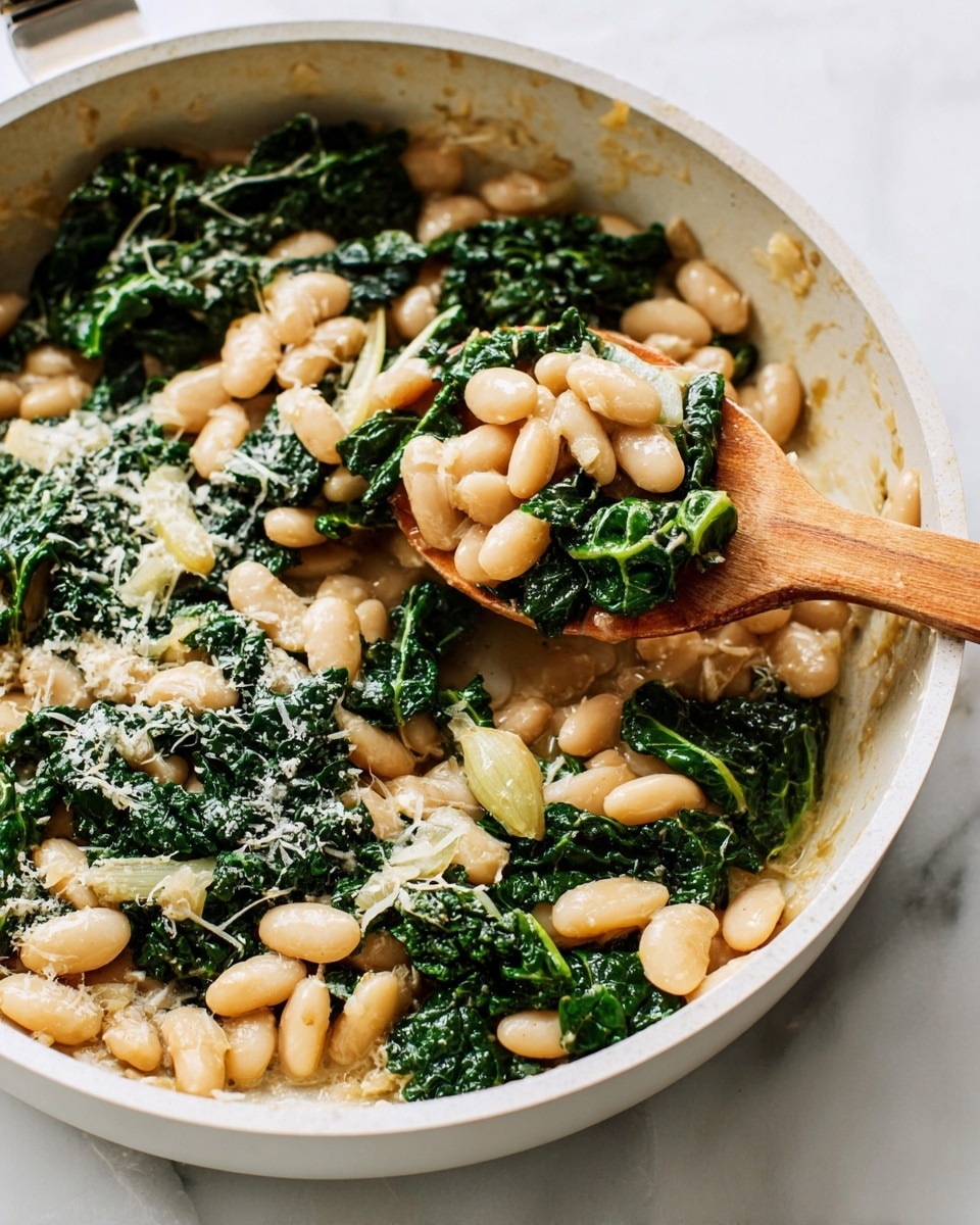 A close-up of a white frying pan filled with cooked white beans and dark green kale leaves mixed together. The beans are light beige, soft, and slightly shiny from cooking, while the kale leaves appear wilted but still hold their dark green color with visible veins. Some light-colored small bits, likely grated cheese or garlic, are sprinkled on top. A wooden spatula is partially lifting a spoonful of the mixture with both beans and kale, showing a creamy texture underneath. The pan sits on a white marbled surface. photo taken with an iphone --ar 4:5 --v 7