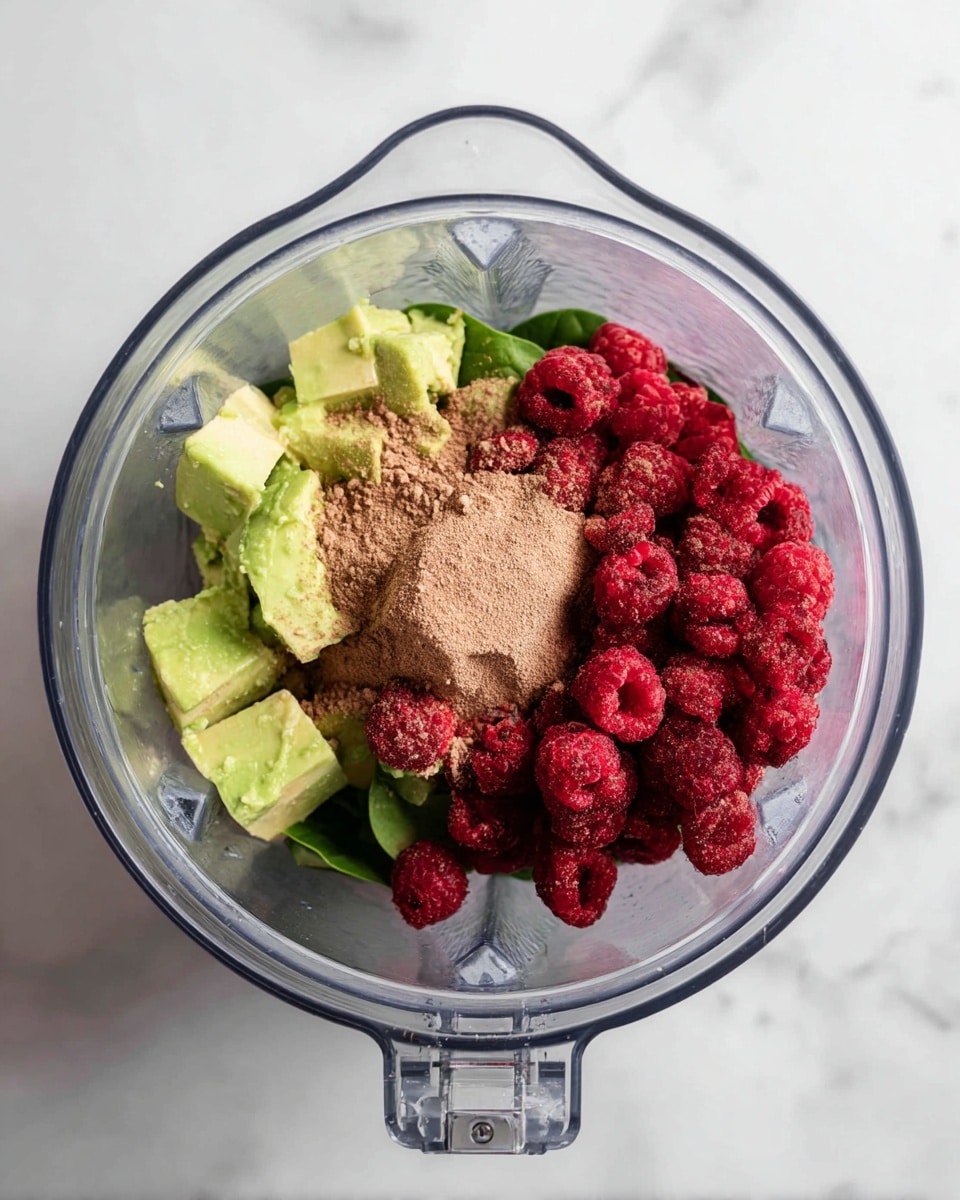 The image shows a clear blender container placed on a white marbled surface, filled with three visible layers of ingredients. The bottom layer is dark green spinach leaves, slightly visible under the other ingredients. On the left side, there are chunks of light green frozen avocado. The right side is covered with a pile of bright red frozen raspberries. On top of the raspberries and avocado is a layer of light brown powder evenly spread across the center. The photo is taken from above, capturing all layers clearly. Photo taken with an iphone --ar 4:5 --v 7