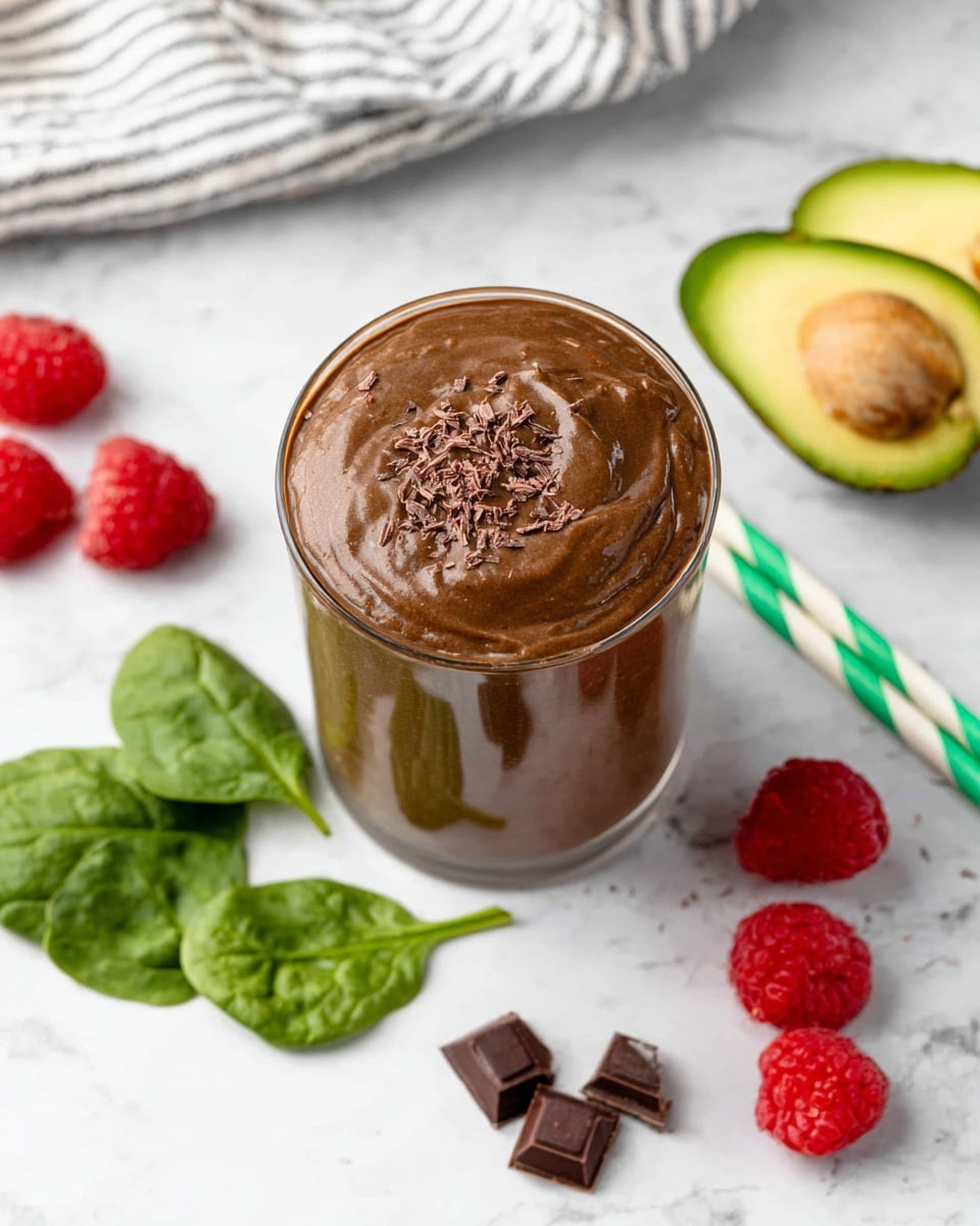 A thick, creamy chocolate smoothie with a shiny texture fills a clear glass, topped with small chocolate shavings. Two white paper straws with green stripes are placed on the right side of the glass, slightly leaning outward. Around the glass on a white marbled surface, there are bright green spinach leaves, fresh red raspberries, dark chocolate chunks, and two halves of a ripe avocado with green skin and light yellow-green flesh. A striped cloth in the background adds a soft texture to the scene. photo taken with an iphone --ar 4:5 --v 7