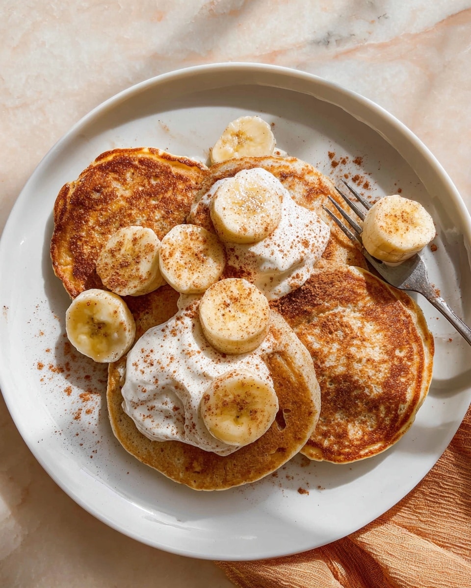 A white plate holds three golden-brown pancakes with slightly crispy edges, arranged with two overlapping at the back and one in front. On top of the pancakes and between the layers are light yellow banana slices sprinkled with brown cinnamon powder. There are three dollops of white whipped cream on the pancakes, also dusted lightly with cinnamon. A silver fork rests on the plate’s bottom right edge, holding a small piece of pancake. The plate is on a white marbled surface with a soft shadow cast around it. Photo taken with an iphone --ar 4:5 --v 7