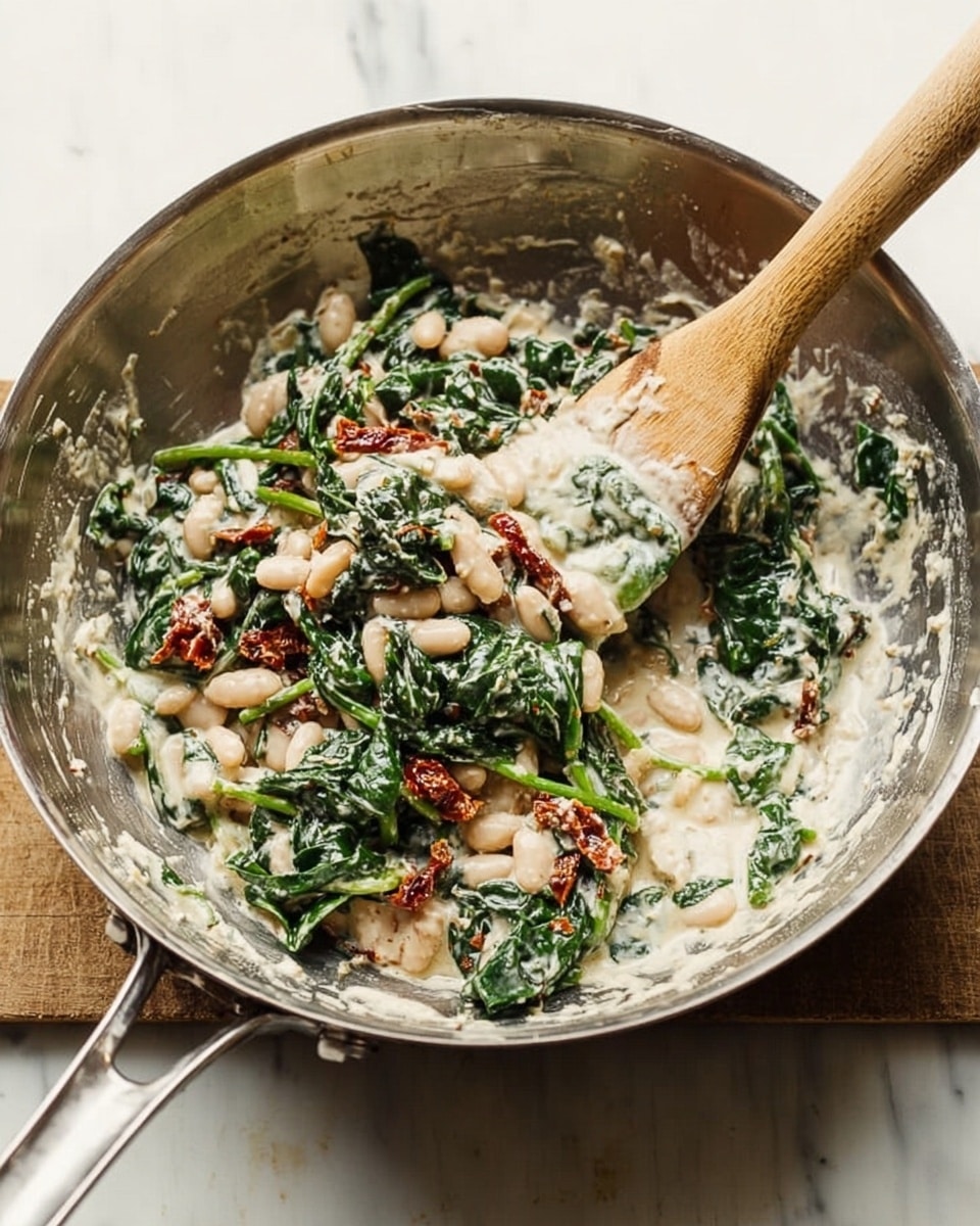 A silver mixing bowl sits on a white marbled surface containing a mixture of white beans, green spinach, and pieces of red sun-dried tomatoes all mixed in a creamy sauce. A wooden spatula with a light-colored handle rests inside the bowl, partially covered with the mixture. The ingredients show varied textures: smooth cream, soft beans, and leafy spinach. Some mixture is spread thinly on the bowl's sides. Photo taken with an iphone --ar 4:5 --v 7