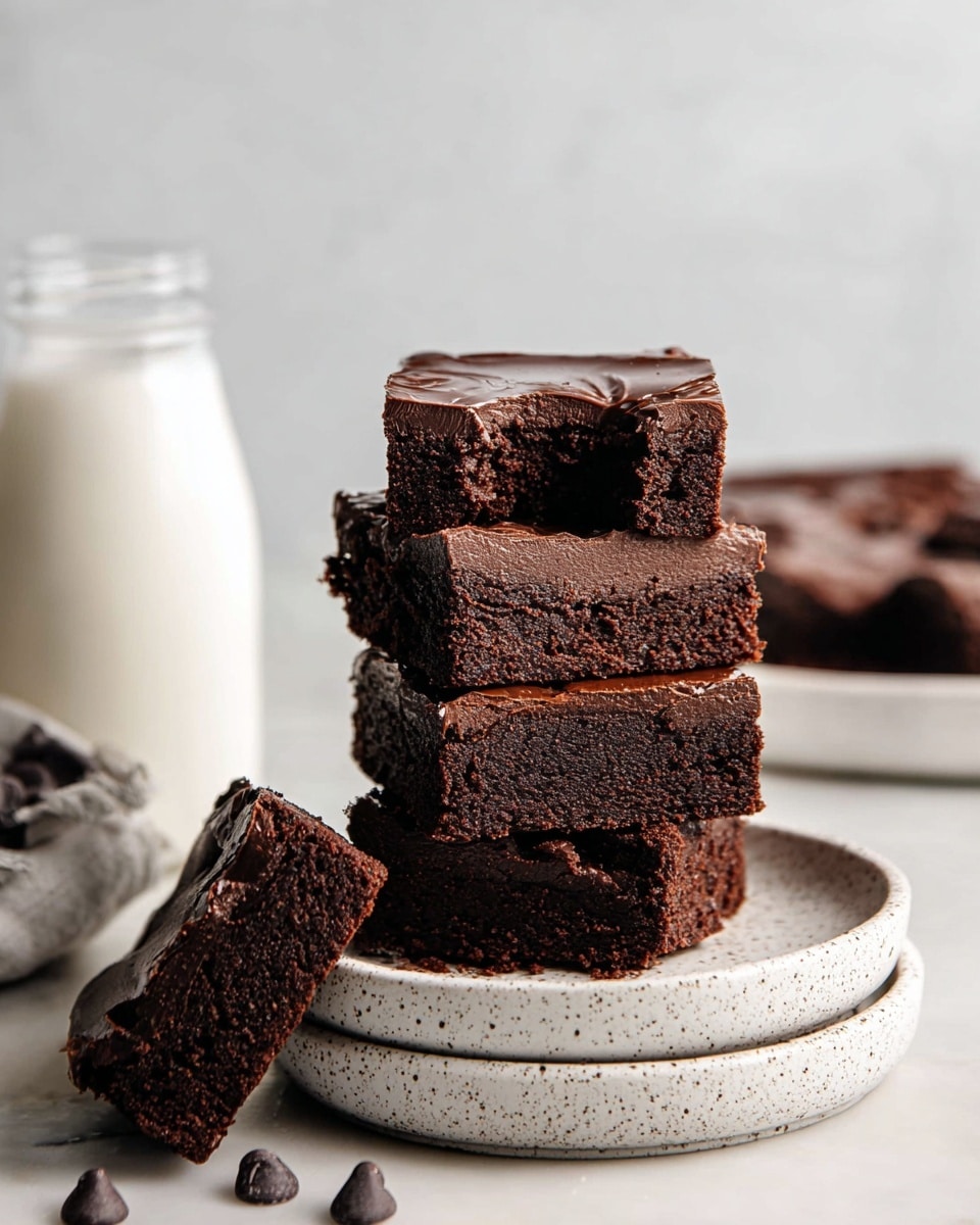 A stack of five thick, dark brown brownies with a smooth, glossy chocolate layer on top sits on a white speckled plate, which is placed on another similar plate. Each brownie is dense and fudgy in texture, with one piece leaning against the stack showing a rich, crumbly interior. The top brownie has a bite taken out of it, revealing the dense inside. Scattered around the plate are several dark chocolate chips. In the background, there is a clear glass bottle filled with white milk, all set on a white marbled surface. Photo taken with an iphone --ar 4:5 --v 7