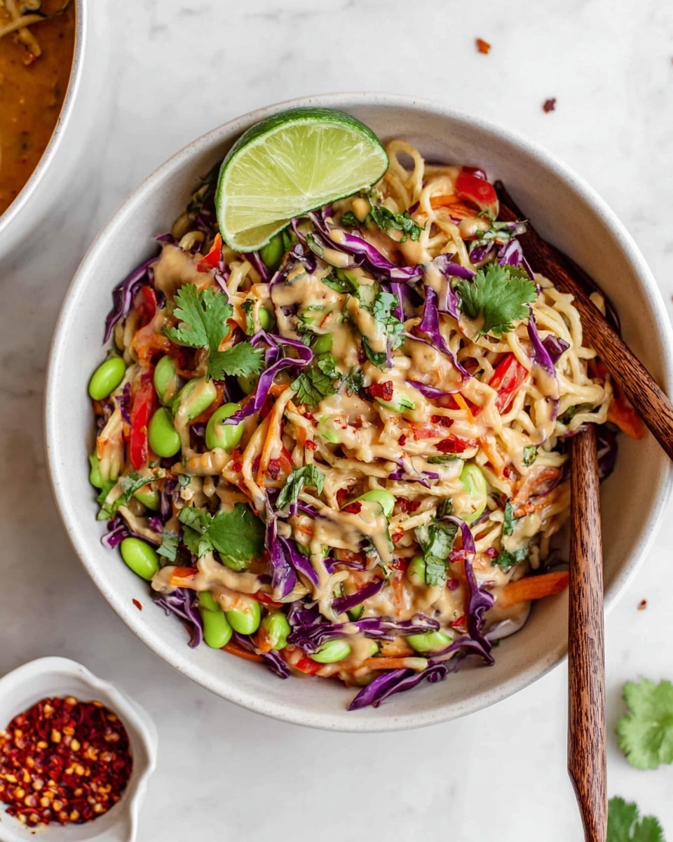 The image shows a white bowl filled with a vibrant noodle salad. The base layer contains light brown noodles mixed with thin strips of purple cabbage and bright green edamame scattered around. On top of this, there are bits of orange carrot and thin slices of red pepper adding more color. A creamy, light brown sauce is drizzled over the dish, with some crushed red chili flakes sprinkled throughout. Fresh green cilantro leaves are spread over the salad, and two wooden chopsticks rest inside the bowl on the right side. A green lime wedge sits on the top left edge of the bowl. The bowl is placed on a white marbled surface with a small white cup of crushed red chili flakes nearby. photo taken with an iphone --ar 4:5 --v 7