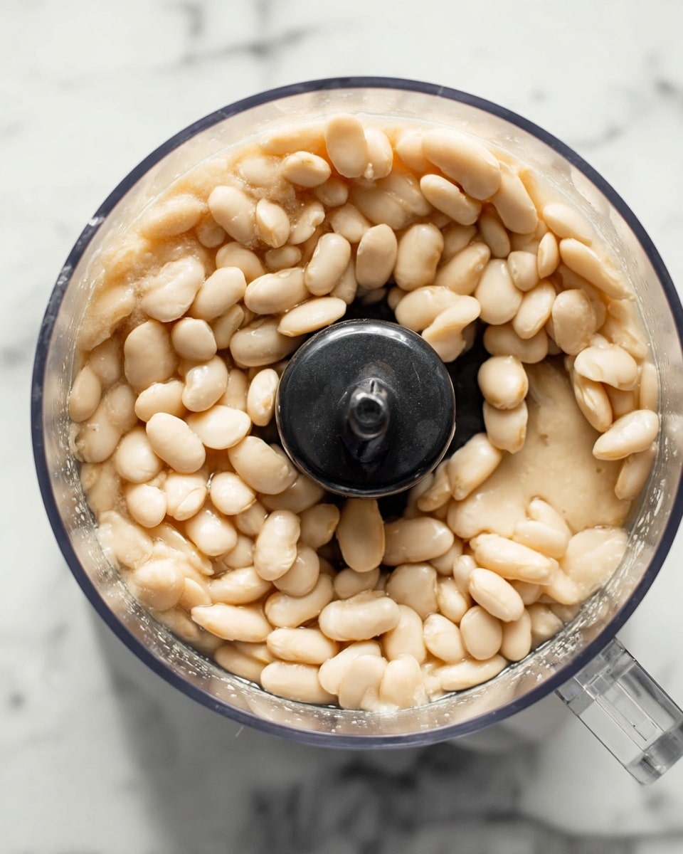 A clear food processor bowl filled with light beige white beans soaking in pale liquid, showing a mix of whole and slightly broken beans with a smooth and soft texture, the black center blade of the food processor visible in the middle, all placed on a white marbled surface photo taken with an iphone --ar 4:5 --v 7