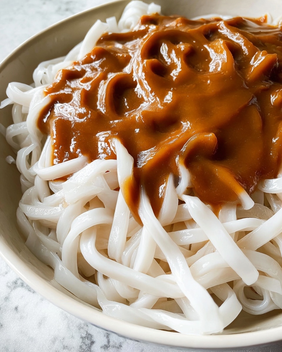 The image shows a close-up view of a bowl filled with thick, white flat noodles that have a smooth and slightly shiny texture. On top of the noodles is a generous layer of thick brown sauce with a glossy surface that looks creamy and rich, slightly dripping over some noodles. The bowl is white, and the background has a white marbled texture. photo taken with an iphone --ar 4:5 --v 7