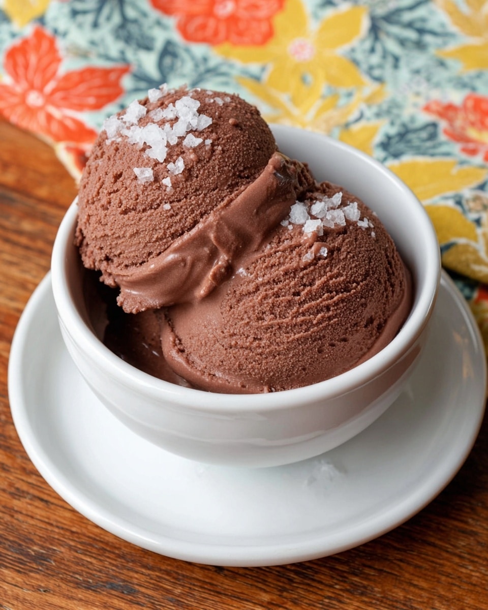 A white bowl filled with two scoops of rich brown chocolate ice cream layered smoothly, slightly melting on one side. The top scoop is sprinkled with coarse white sea salt crystals that add texture. The bowl sits on a white plate on a wooden table with a colorful floral cloth in the background featuring orange, red, yellow, and green hues. Photo taken with an iphone --ar 4:5 --v 7