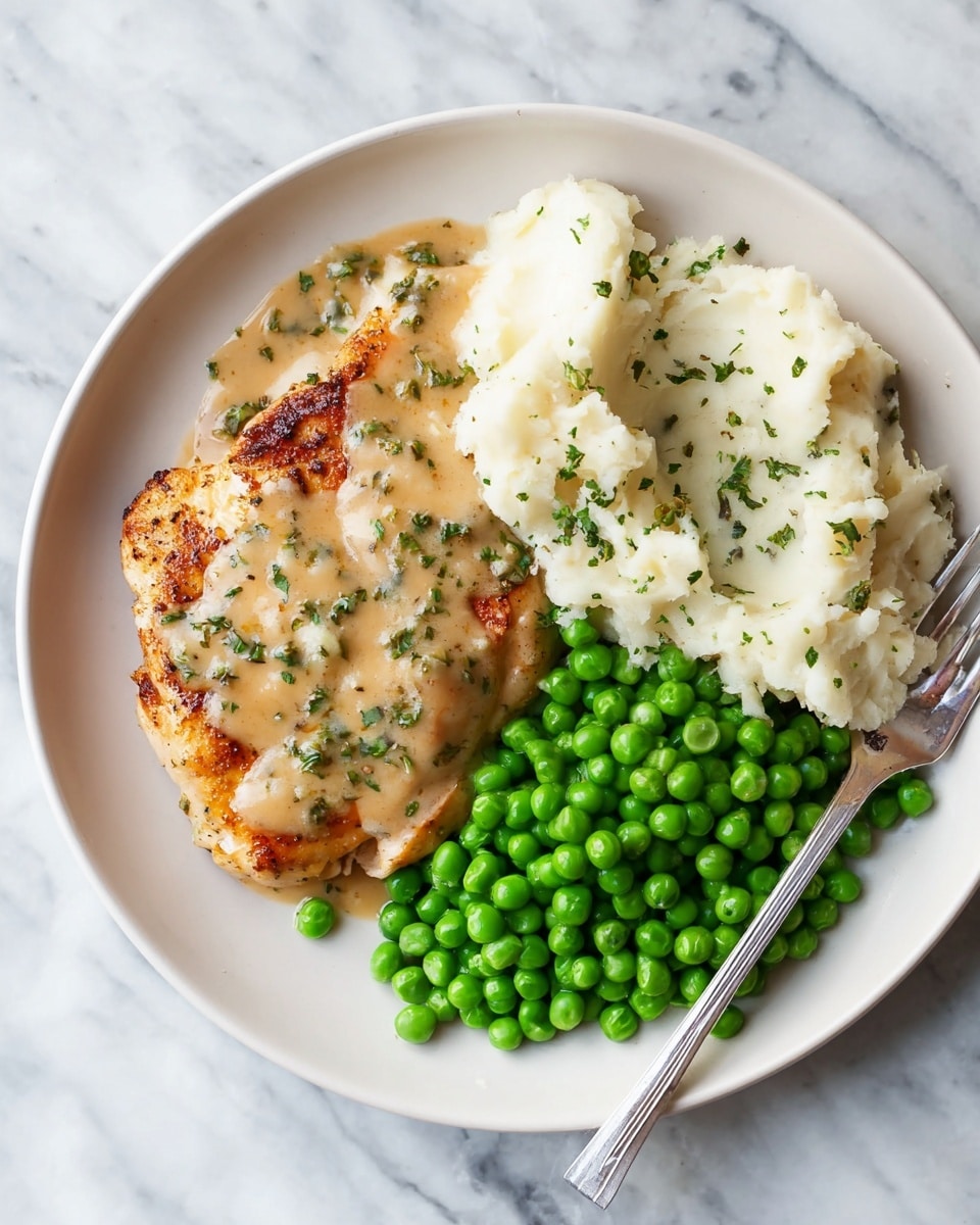 The image shows a white plate placed on a white marbled surface, filled with three main layers of food. On the left side, there is a browned grilled chicken piece topped with a light brown creamy sauce with tiny green herb bits. In the middle top section of the plate, there are fluffy white mashed potatoes covered partially by the same creamy sauce. On the right side, a pile of bright green peas fills the space. A silver fork is placed on the right edge of the plate with some mashed potatoes and sauce near it. Photo taken with an iphone --ar 4:5 --v 7