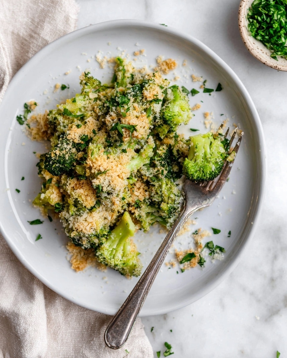On a white plate, there is a serving of broccoli topped with a crumbly layer of light brown and off-white breadcrumbs mixed with green herbs. The broccoli pieces are bright green and placed in a small pile, with some crumbs scattered on the plate. A silver fork rests on the right side of the plate with a small broccoli floret speared on its tines. The plate is set on a white marbled surface, and there is a small bowl with chopped green herbs visible in the top right corner. Photo taken with an iphone --ar 4:5 --v 7