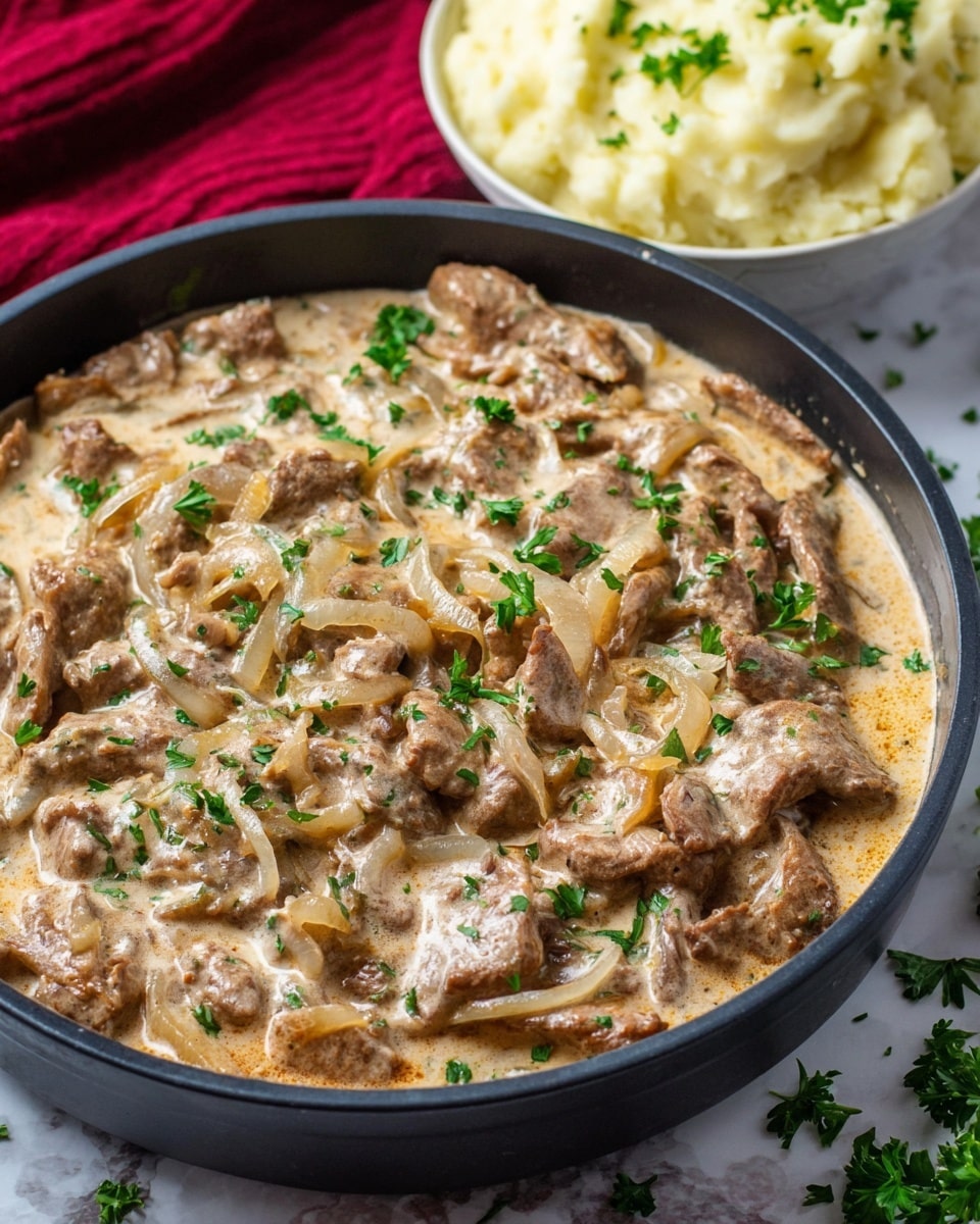 A close-up shows a black pan filled with creamy beige sauce layered with light brown, thin strips of cooked meat and translucent sautéed onions. Bright green parsley leaves are scattered on top, adding fresh color. In the background, there is a white bowl filled with soft, pale yellow mashed potatoes sitting on a red cloth, all placed on a white marbled surface with a few parsley leaves sprinkled around. photo taken with an iphone --ar 4:5 --v 7