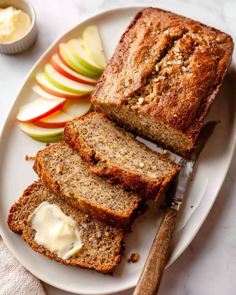 A loaf of brown bread with a crispy crust sits on a white oval plate on a white marbled surface. Four thick slices are cut and laid flat in front of the loaf, showing a soft, moist inside with small bits of nuts or grains. The closest slice is spread with a light layer of butter. Next to the slices on the plate are thinly sliced red and green apples arranged in small stacks. A small round container of butter and a butter knife with a wooden handle rest on the plate near the bread. Photo taken with an iphone --ar 4:5 --v 7