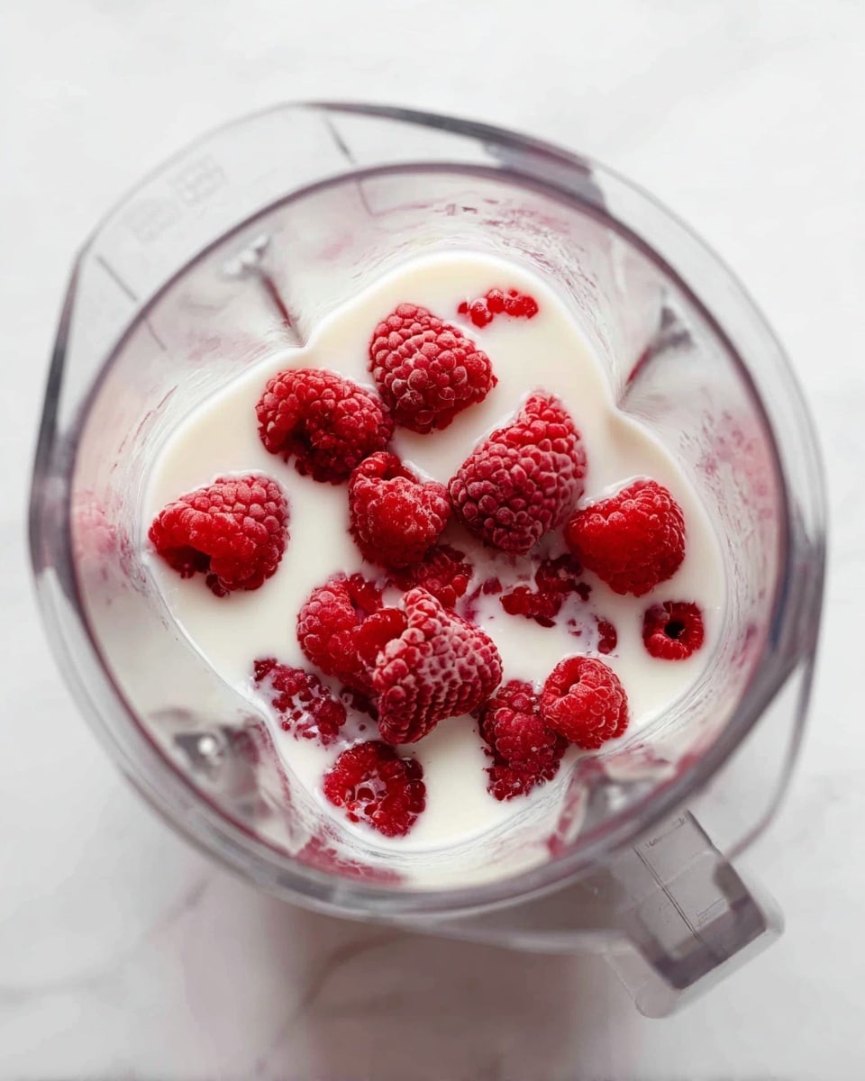 A clear blender container shows fresh red raspberries sitting partially submerged in white milk, with some raspberries floating on the milk's surface and others at the bottom. The raspberries have a textured, bumpy surface and deep red color, contrasting with the smooth, creamy white milk. The blender container has angled, clear walls that reflect light softly, and the entire setup is placed on a white marbled surface. photo taken with an iphone --ar 4:5 --v 7