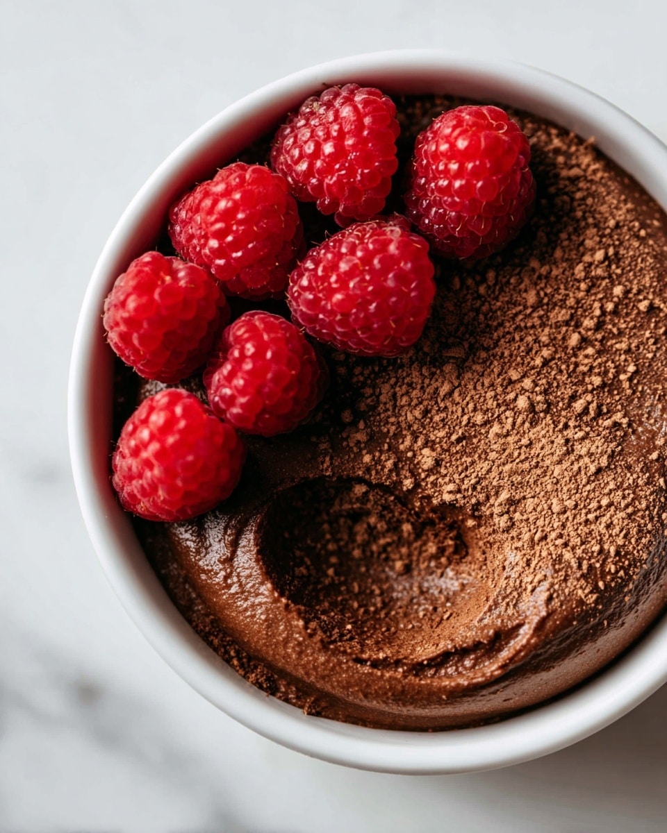 A close-up view of a white bowl filled with a single layer of rich, dark brown chocolate mousse with a soft, slightly bumpy texture. The mousse has a small round indentation on one side revealing a smoother, shiny surface underneath. On top of the mousse, there are five bright red raspberries arranged in a loose cluster near the lower left edge of the bowl. There is a light dusting of cocoa powder scattered over the mousse around the raspberries. The bowl sits on a white marbled surface. photo taken with an iphone --ar 4:5 --v 7