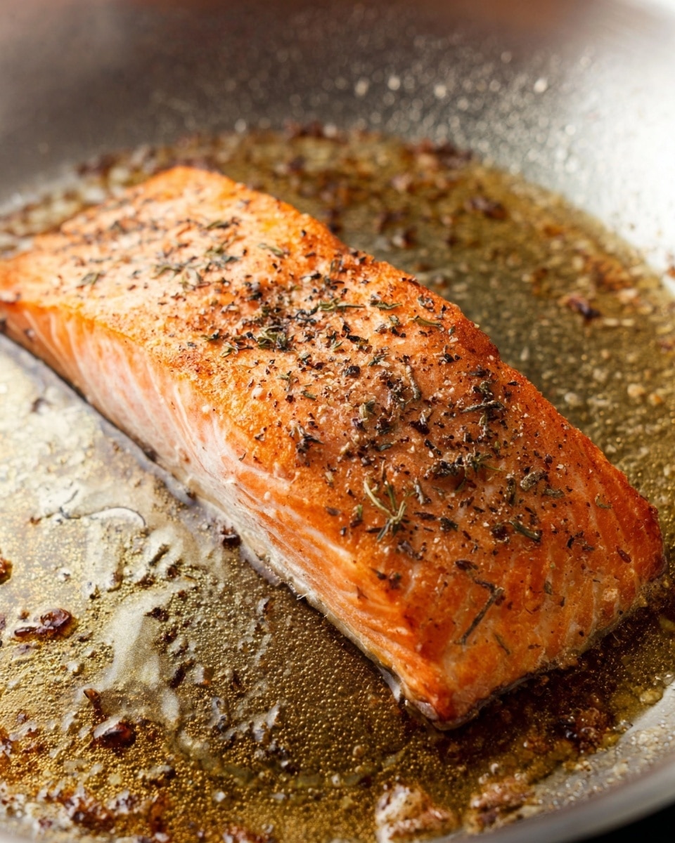 A single thick piece of salmon fillet is shown cooking in a stainless steel pan. The salmon has a light orange-pink color with crispy, lightly browned edges and is sprinkled with black pepper and dried herbs. The surface of the fillet shows a slightly flaky texture. The pan has sizzling oil around the salmon with some browned bits stuck to the pan, giving a warm, cooked look. The background shows no plate but the pan's metallic texture. Photo taken with an iphone --ar 4:5 --v 7