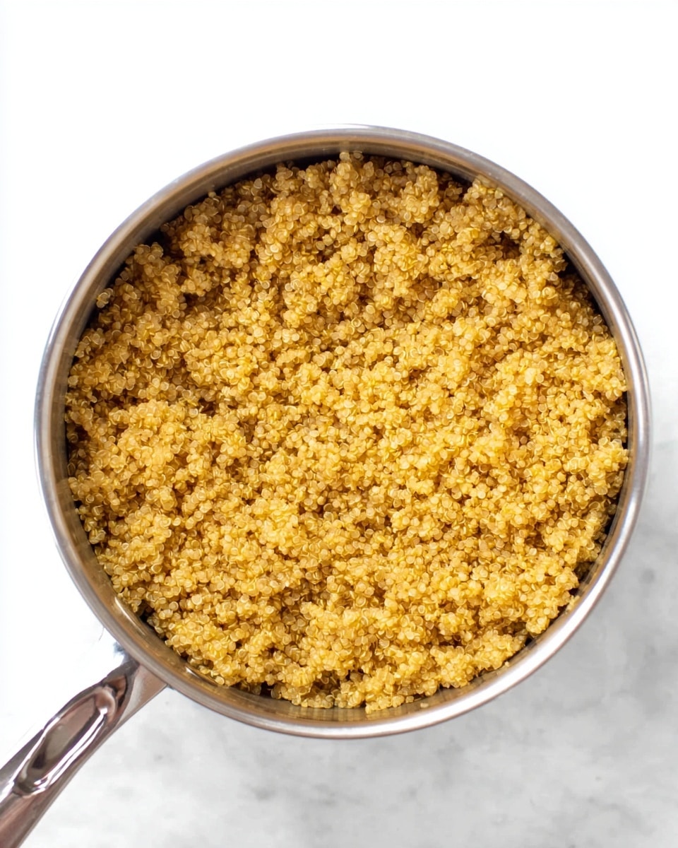 The image shows a single layer of cooked quinoa inside a silver pot, filling the pot almost to the top. The quinoa is light golden brown with a slightly grainy texture, appearing fluffy and evenly cooked. The pot has a shiny metal handle on the bottom left and is placed against a white marbled surface, which is mostly out of focus. Photo taken with an iphone --ar 4:5 --v 7