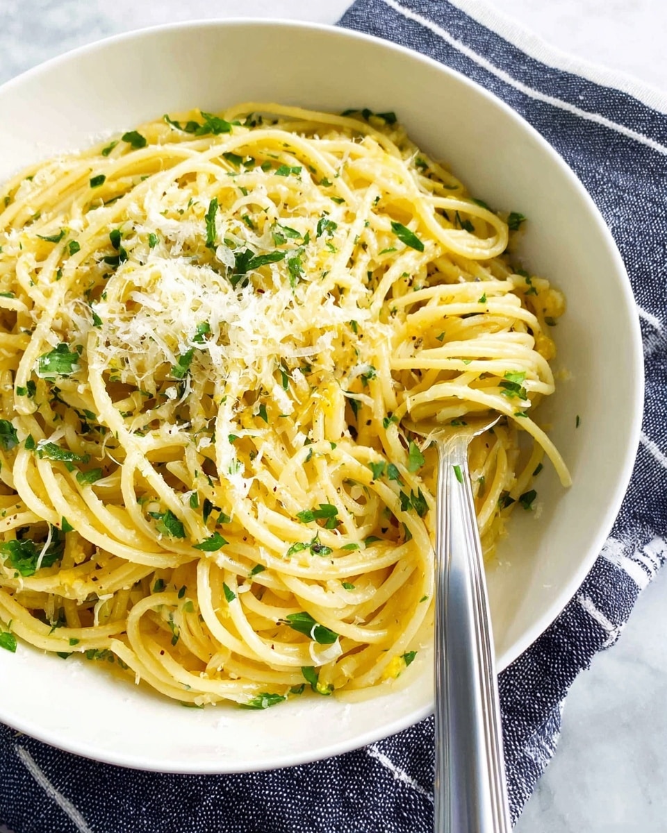 A white bowl filled with twisted spaghetti noodles showing a shiny, light yellow color, mixed with small pieces of green herbs scattered throughout. On top of the noodles, there is a sprinkle of grated pale yellow cheese creating a soft textured layer. A silver fork is resting inside the bowl on the right side, twirling some noodles around its tines. The bowl is placed on a dark blue and white striped cloth on a white marbled surface. photo taken with an iphone --ar 4:5 --v 7