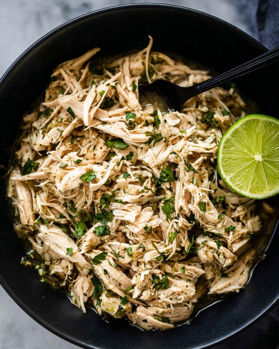 A close-up view of a black bowl filled with shredded light brown chicken mixed with small pieces of green herbs, giving it a fresh look. A wedge of bright green lime sits on the right side inside the bowl. A black spoon is partially resting in the chicken. The chicken looks moist with some sauce visible at the bottom of the bowl. The bowl is placed on a white marbled surface. Photo taken with an iphone --ar 4:5 --v 7