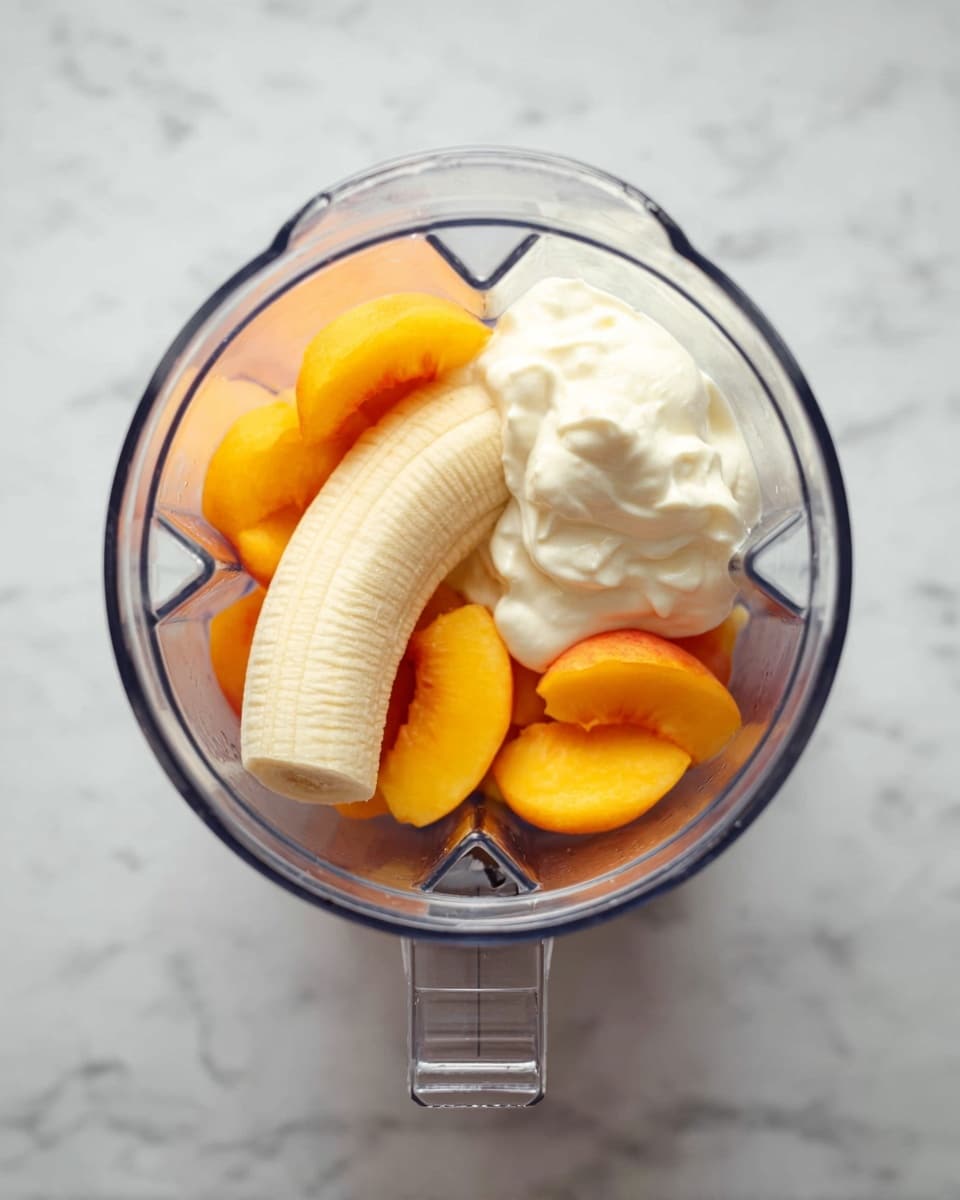 A clear blender container on a white marbled surface holds three main ingredients in separate layers. At the bottom, there are bright orange peach slices arranged mostly in the center. On the left side, a whole peeled banana with a smooth pale yellow color rests. On the right side, a dollop of thick, creamy white yogurt with a slightly uneven texture sits next to the fruit. The blender is viewed from above, showing the clear blades at the base inside the container. photo taken with an iphone --ar 4:5 --v 7