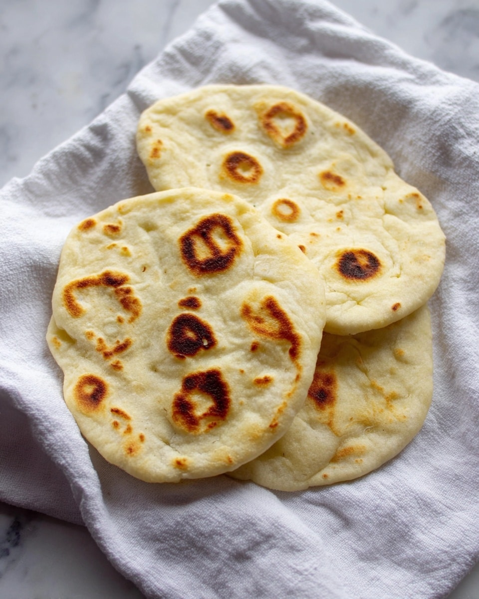 The image shows three flatbreads with a soft and fluffy texture, each having a light cream color with some brown toasted spots. They are stacked casually on top of a white cloth with wrinkles, placed on a white marbled surface. The flatbreads are round but slightly uneven in shape, and the toasted spots vary in size and placement on each piece. photo taken with an iphone --ar 4:5 --v 7