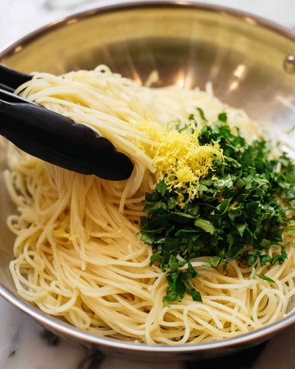 Thin spaghetti pasta forms the base layer, showing a pale yellow color with a soft and smooth texture. On top of the noodles, there is a small pile of fine yellow lemon zest, adding a bright touch on the right side. Above the lemon zest, chopped fresh green herbs cover part of the pasta, with varying shades of green and a leafy, uneven texture. A pair of black kitchen tongs hold and lift some pasta and herbs on the left side. The dish is in a large pan with a metallic surface as the background, though the setting is changed to a white marbled texture. Photo taken with an iphone --ar 4:5 --v 7