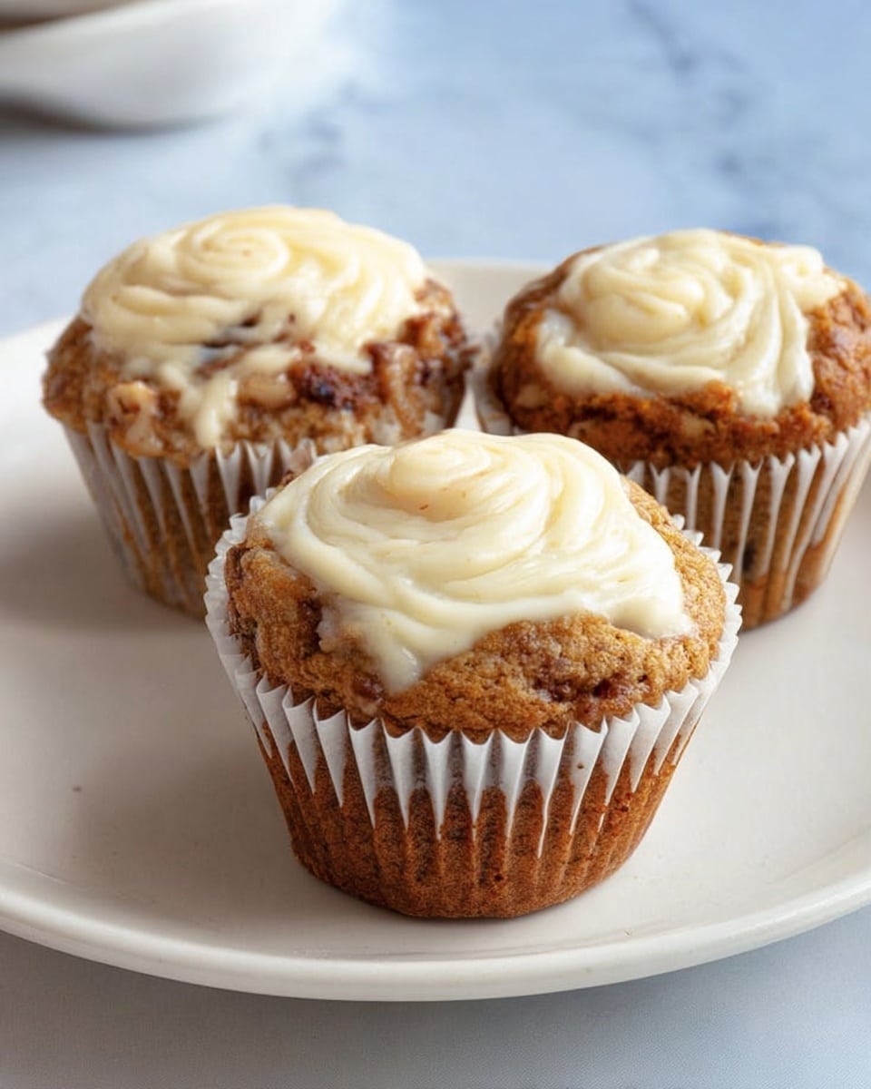 The image shows three muffins with a swirl of creamy white topping on each. The muffins are golden brown with visible darker spots of nuts or spices inside. They sit on a smooth white plate placed on a white marbled surface. The muffins have white paper liners, and the creamy topping creates a soft, textured pattern on the top of each muffin. Photo taken with an iphone --ar 4:5 --v 7