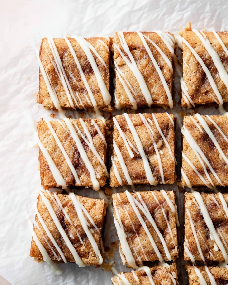 The image shows nine square pieces of a light brown baked dessert arranged in a three-by-three grid on white parchment paper over a white marbled surface. Each piece has a slightly rough, cinnamon-speckled texture on top. All squares are drizzled with thin, white icing lines, applied unevenly but consistently across the pieces. The dessert looks soft with some darker cinnamon swirls visible just below the surface. Photo taken with an iphone --ar 4:5 --v 7