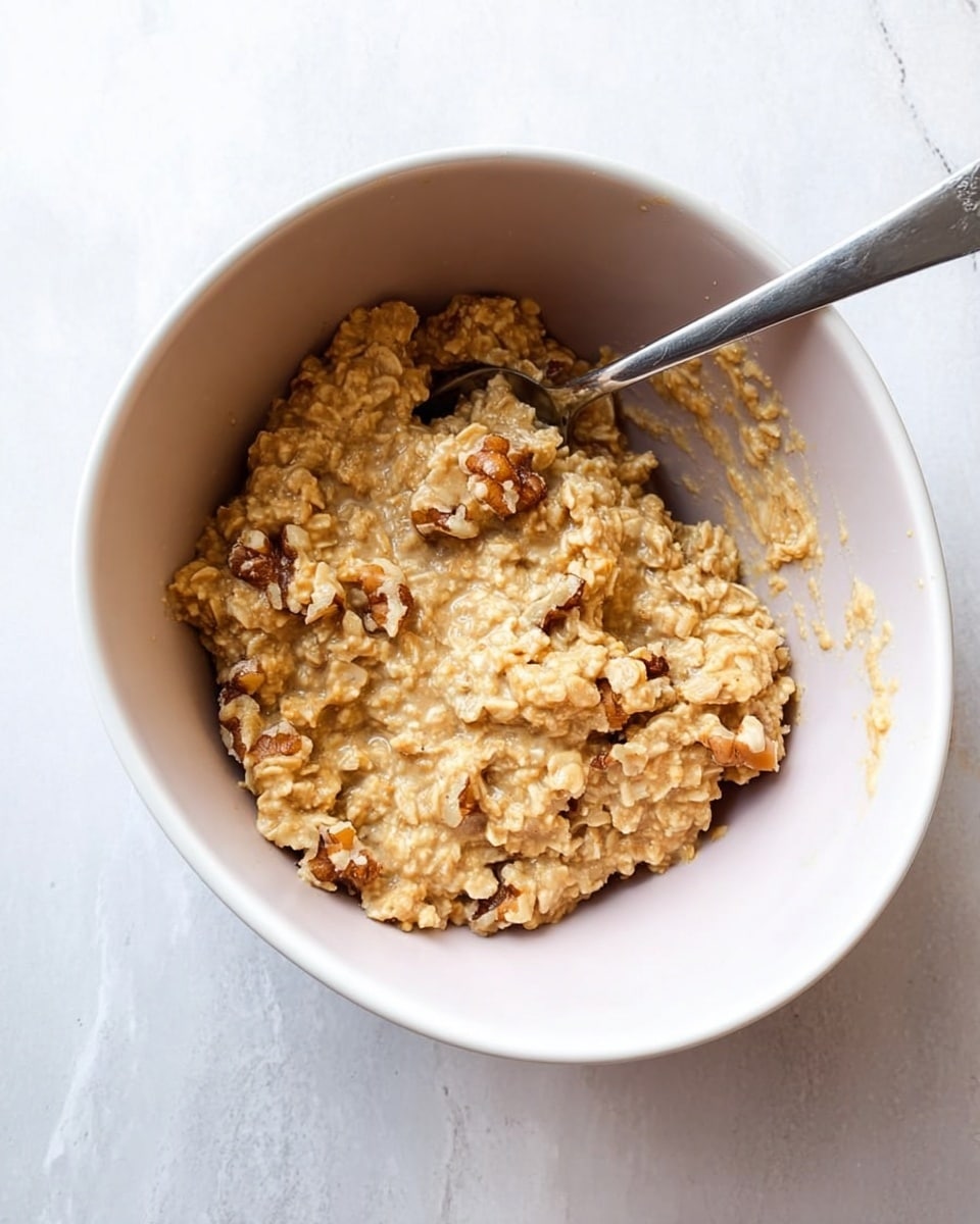 The image shows a white bowl filled with a thick, light brown oatmeal mixture with visible pieces of walnuts mixed in. A silver spoon is placed inside the bowl, resting against the curved inner wall. The oatmeal looks soft and somewhat chunky with an uneven texture. The background is a white marbled surface, giving a clean and bright look to the scene. Photo taken with an iphone --ar 4:5 --v 7