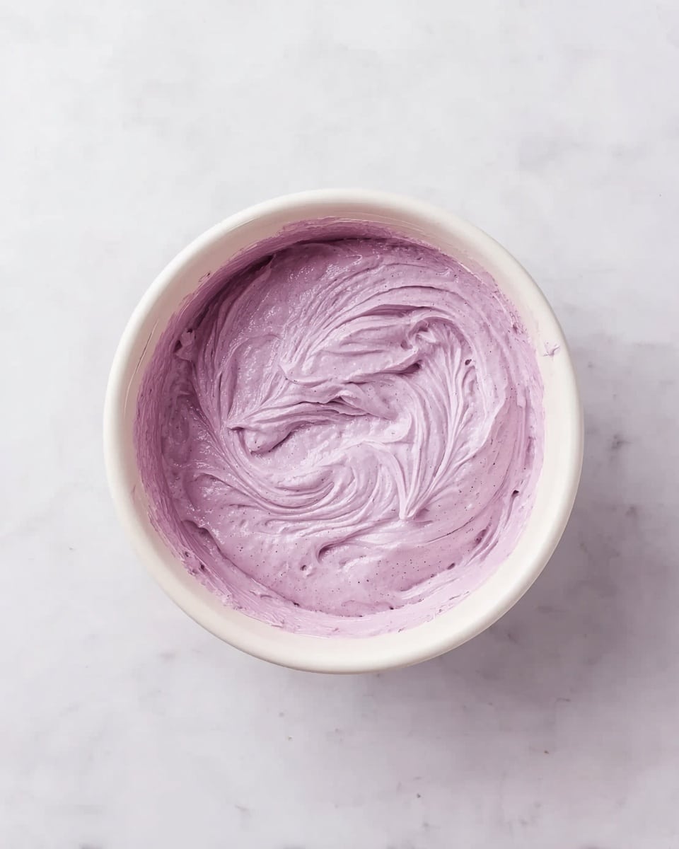 A top view of a single white round bowl filled with a creamy, thick mixture of pale purple color. The mixture has soft swirls and a smooth texture that shows slight waves and peaks, indicating it has been stirred recently. The bowl sits on a surface with a white marbled texture. Photo taken with an iphone --ar 4:5 --v 7